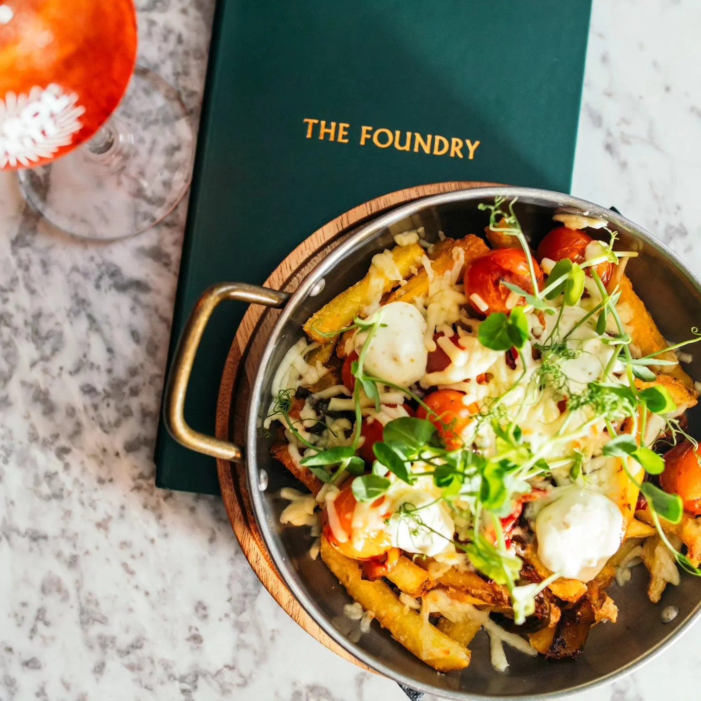 A colorful dish of roasted vegetables topped with microgreens, served in a small metal pan with a handle, on a wooden board. A green menu labeled 'The Foundry' is underneath the dish, and a glass of rose wine with a white floral design is partially visible in the background on a speckled granite countertop.
