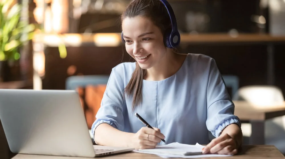 Young woman wearing headphones, smiling, working on a laptop and taking notes in a coffee shop or coworking space.
