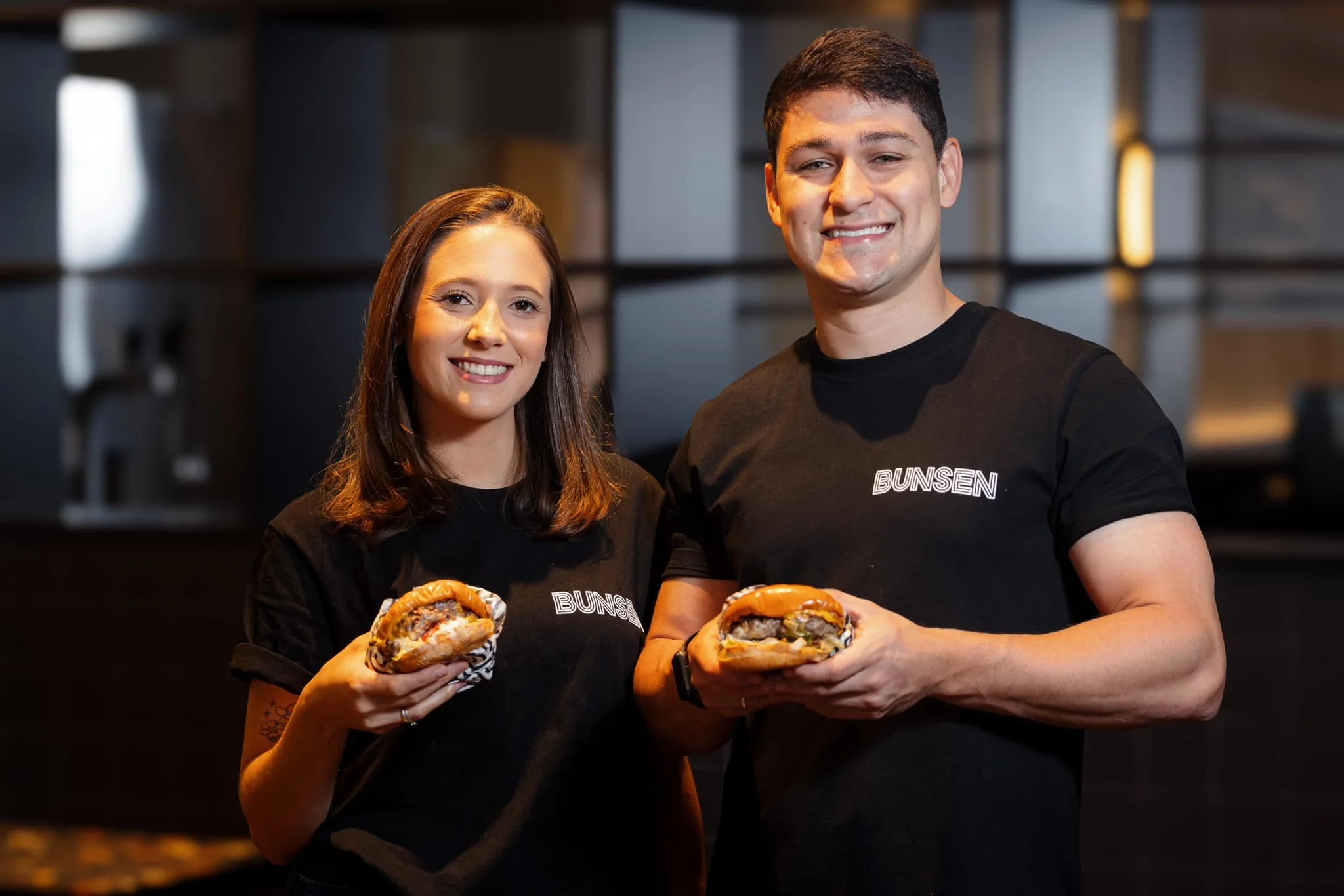 Two young adults, a woman and a man, smiling and holding sandwiches or burgers, standing in a modern indoor setting with dark background and industrial-style shelving.