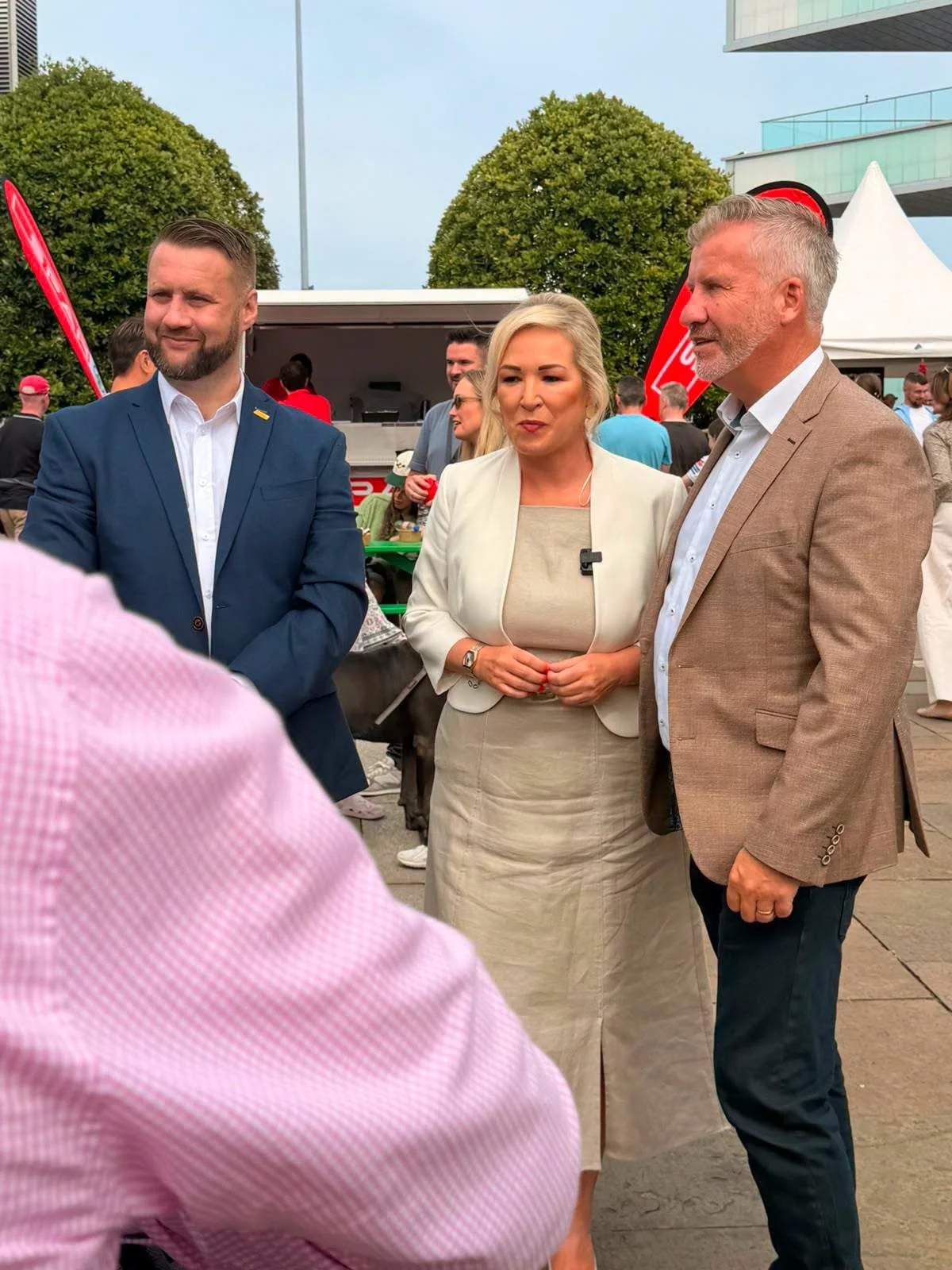 Three people engaged in conversation at an outdoor event. The woman in the middle is wearing a cream blazer and dress, holding her hands together. The man on her right has gray hair, a beard, and is dressed in a tan blazer with a light-colored shirt. The man on her left has short hair, a beard, and is wearing a blue suit. There are other attendees and tents visible in the background.
