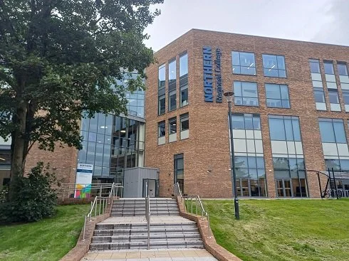 Modern brick and glass building with a sign reading 'NORTHWEST' and 'HEALTH EDUCATION' on the side, with stairs leading up to the entrance, surrounded by trees and a lamppost.