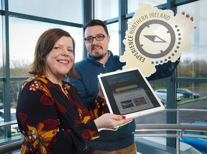 Two people, a woman with shoulder-length brown hair wearing a floral blouse, and a man with glasses and a blue sweater, standing inside a modern building with large glass windows. The woman is holding a tablet displaying a website, and the man holds up a large cutout sign of Northern Ireland with a compass and the text 'Experience Northern Ireland.'