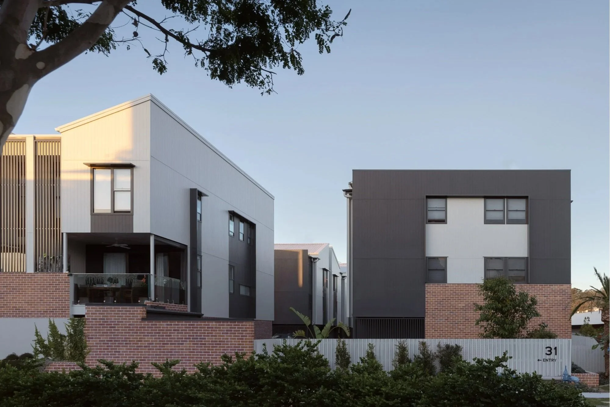 Modern multi-story residential buildings with white and dark gray exterior walls, featuring large windows, situated behind a brick wall at the entrance of a neighborhood, with a blue sky and trees in the background.