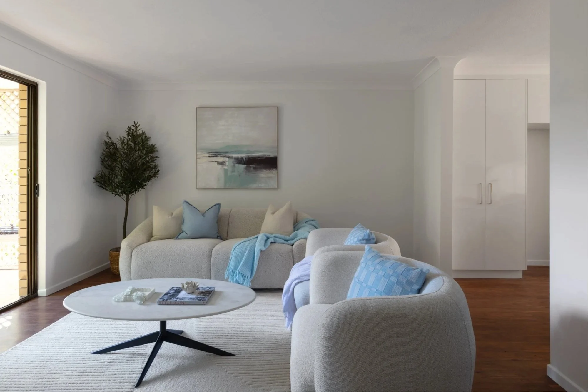Living room with white carpet, beige couches with blue and white pillows, a black coffee table with magazines, a potted plant, and artwork on the wall. Part of a sliding door and white cabinets are visible.