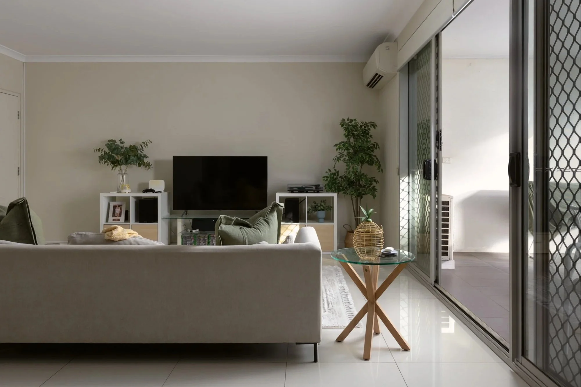 Living room with beige walls, white shelving unit, black TV, green plants, glass coffee table, and sliding glass door with security screen.