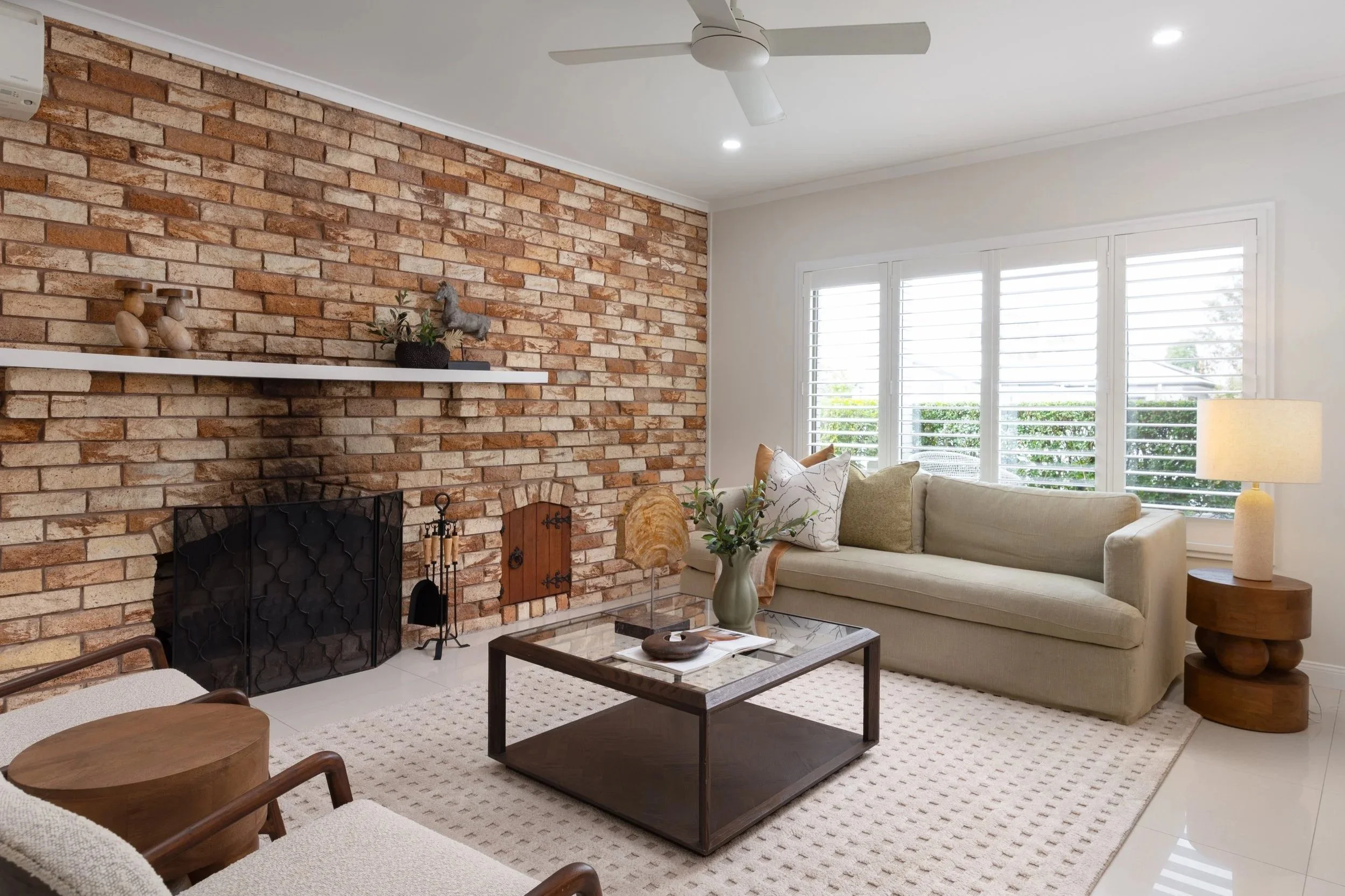Living room with a brick fireplace, white ceiling fan, large window with white blinds, beige sofa with cushions, wooden armchairs, coffee table with decorative items, and a beige area rug.