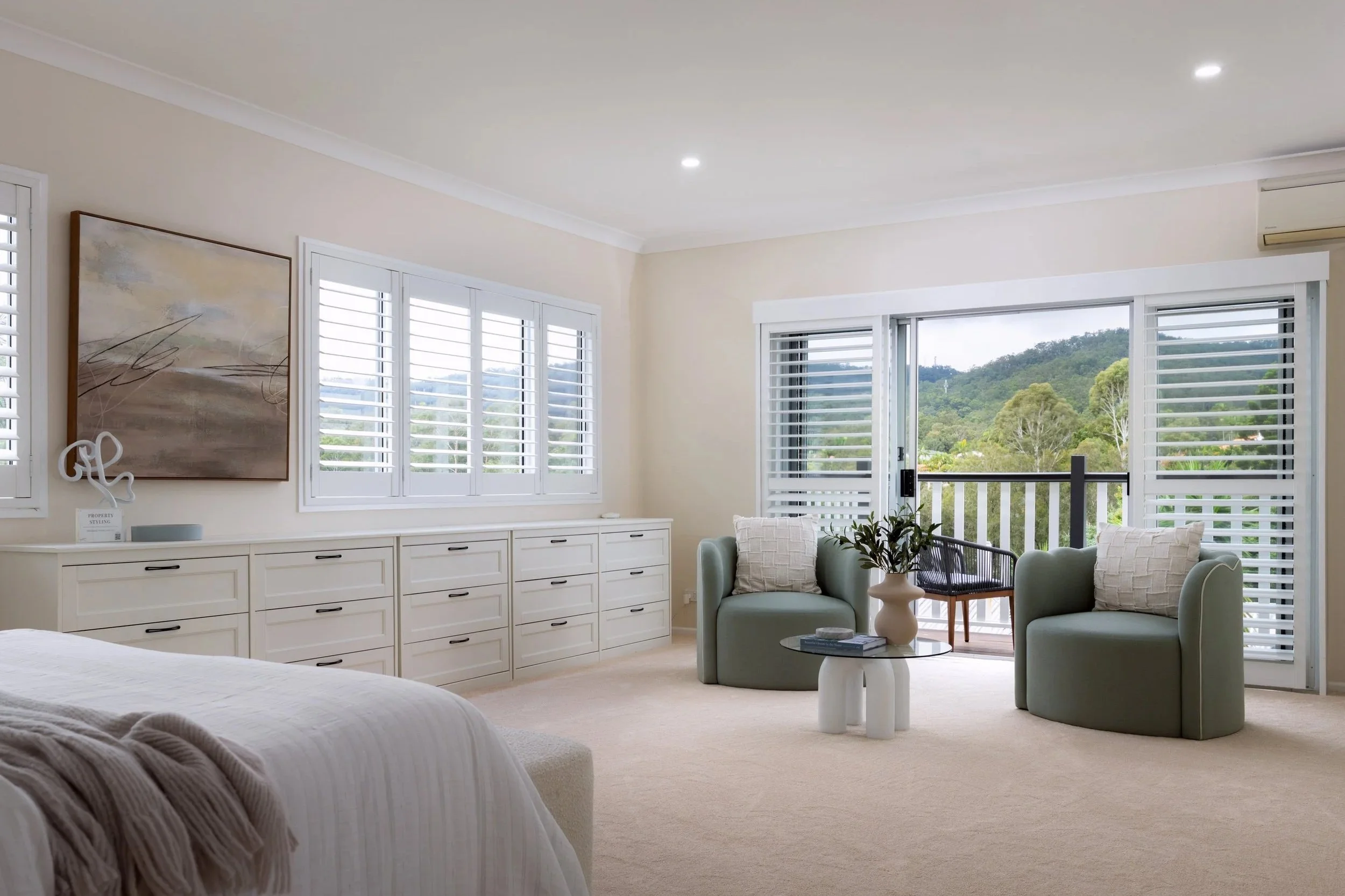 A bright bedroom with large windows overlooking a green, hilly landscape. The room features white furniture, including a dresser and side tables, and two light green armchairs with pillows. There is a modern abstract painting on the wall and a slidin