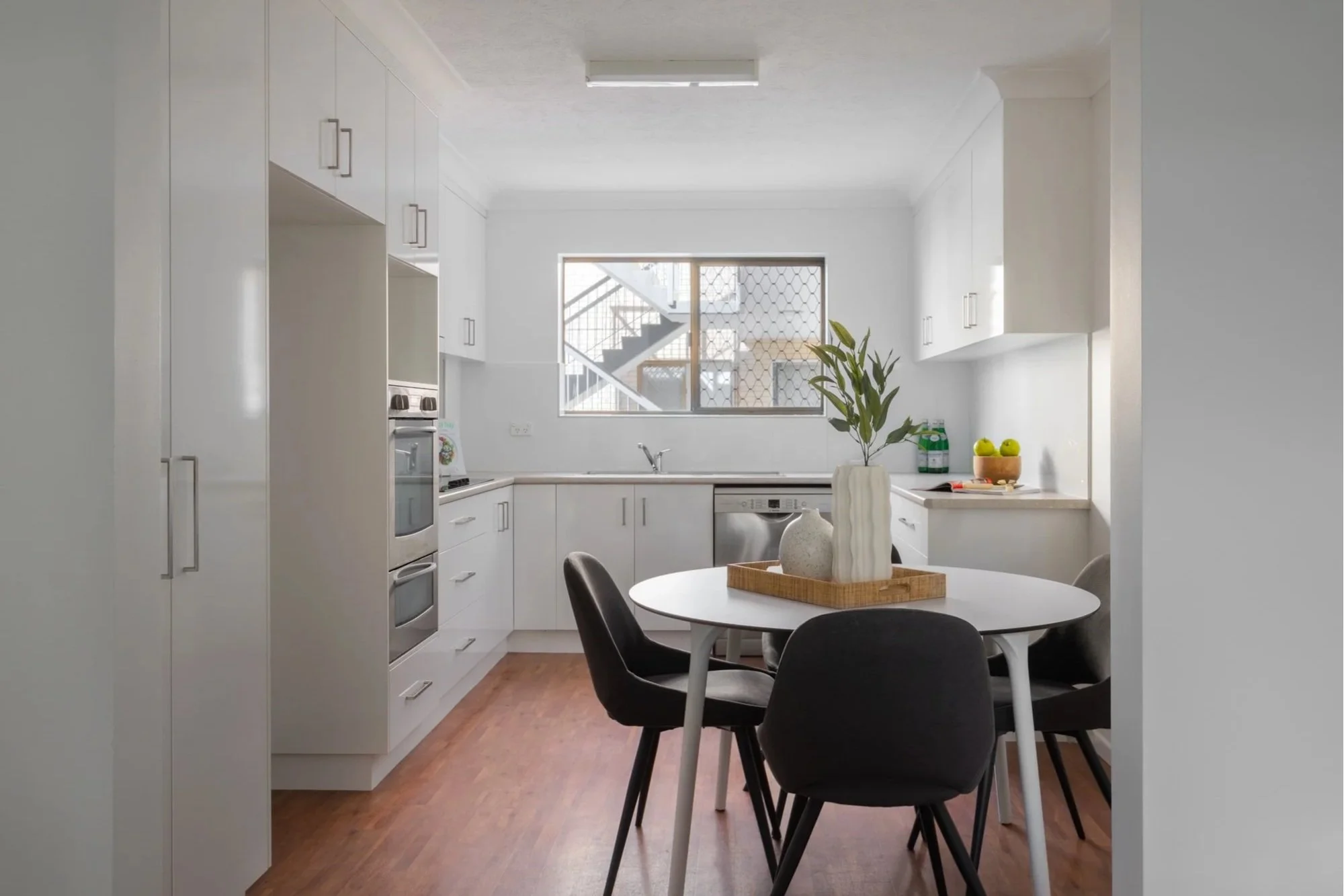 A modern kitchen with white cabinets and a round dining table with five black and white chairs. There is a potted plant and some apples on the table, and a window showing outdoor stairs and building exterior.