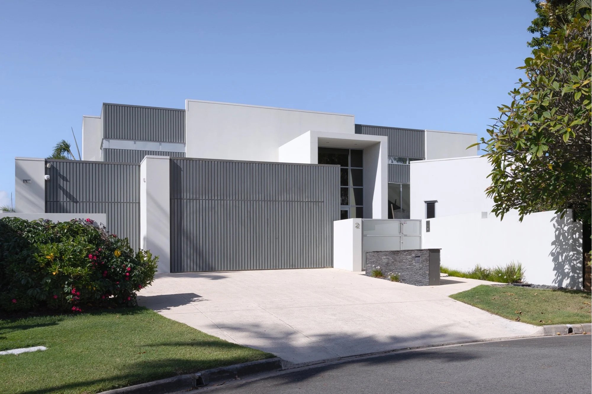 Modern white house with black accents, large windows, and a gated entrance, surrounded by greenery and located along a sidewalk on a sunny day.