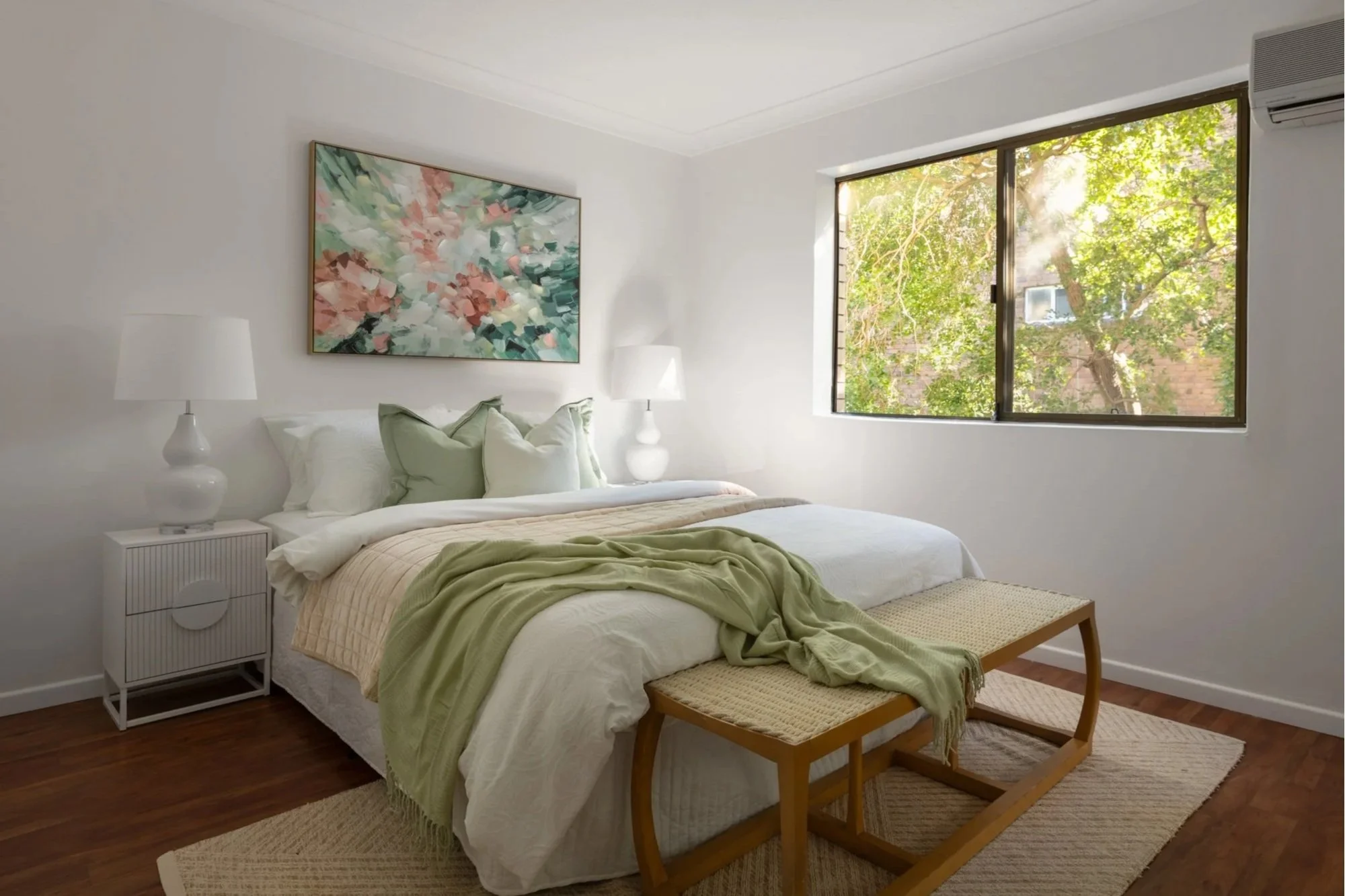 A bedroom with a white bed, green blanket, and multiple pillows. There is a wooden bench at the foot of the bed. White nightstands with lamps on each side of the bed. Large window showing green trees outside. Artwork with abstract floral design hangi