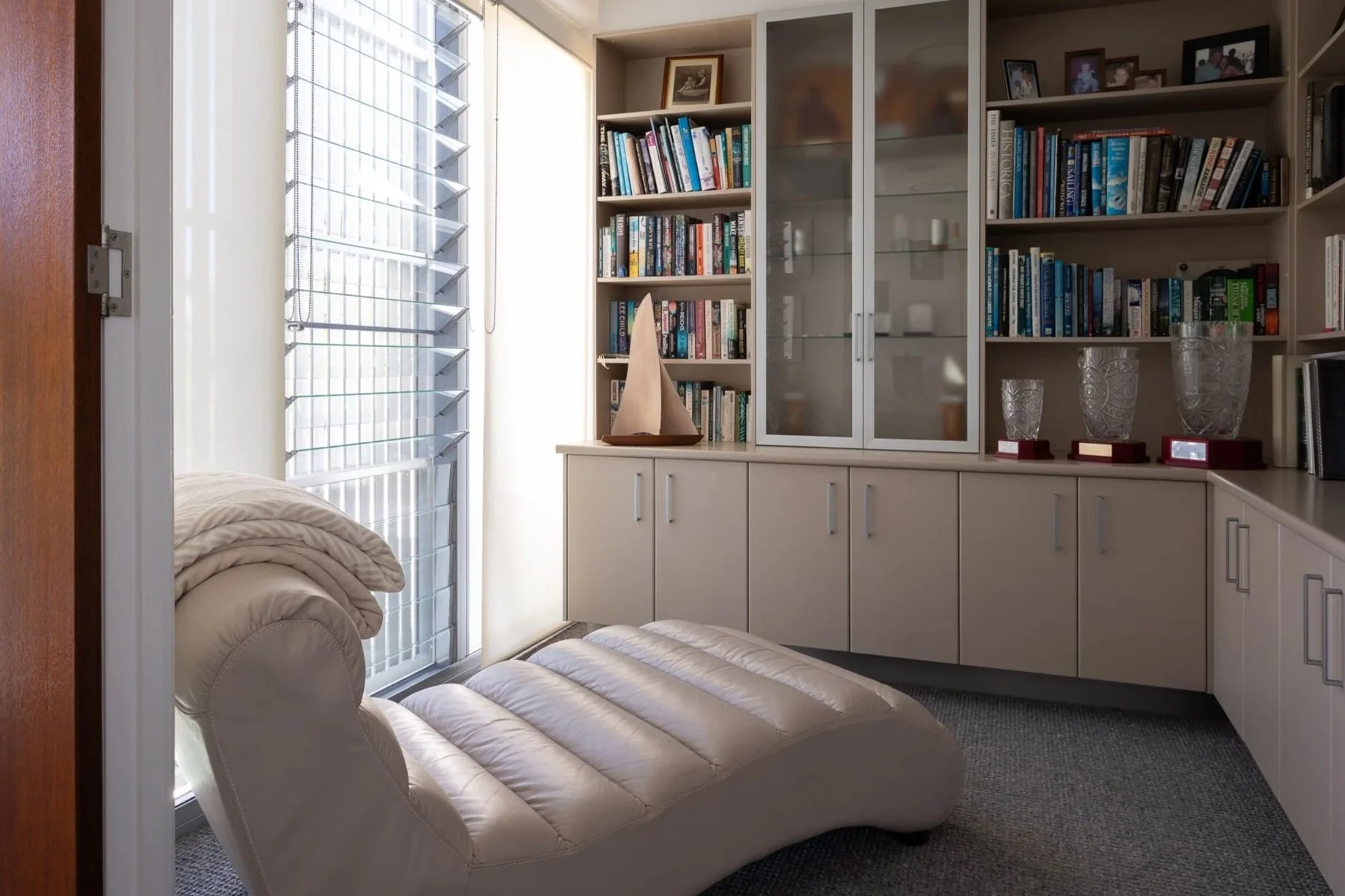 A cozy living room corner with a beige leather futon, a large window with white blinds, a white bookshelf filled with books and framed photos, and a cabinet with glass doors displaying trophies and decorative glassware.