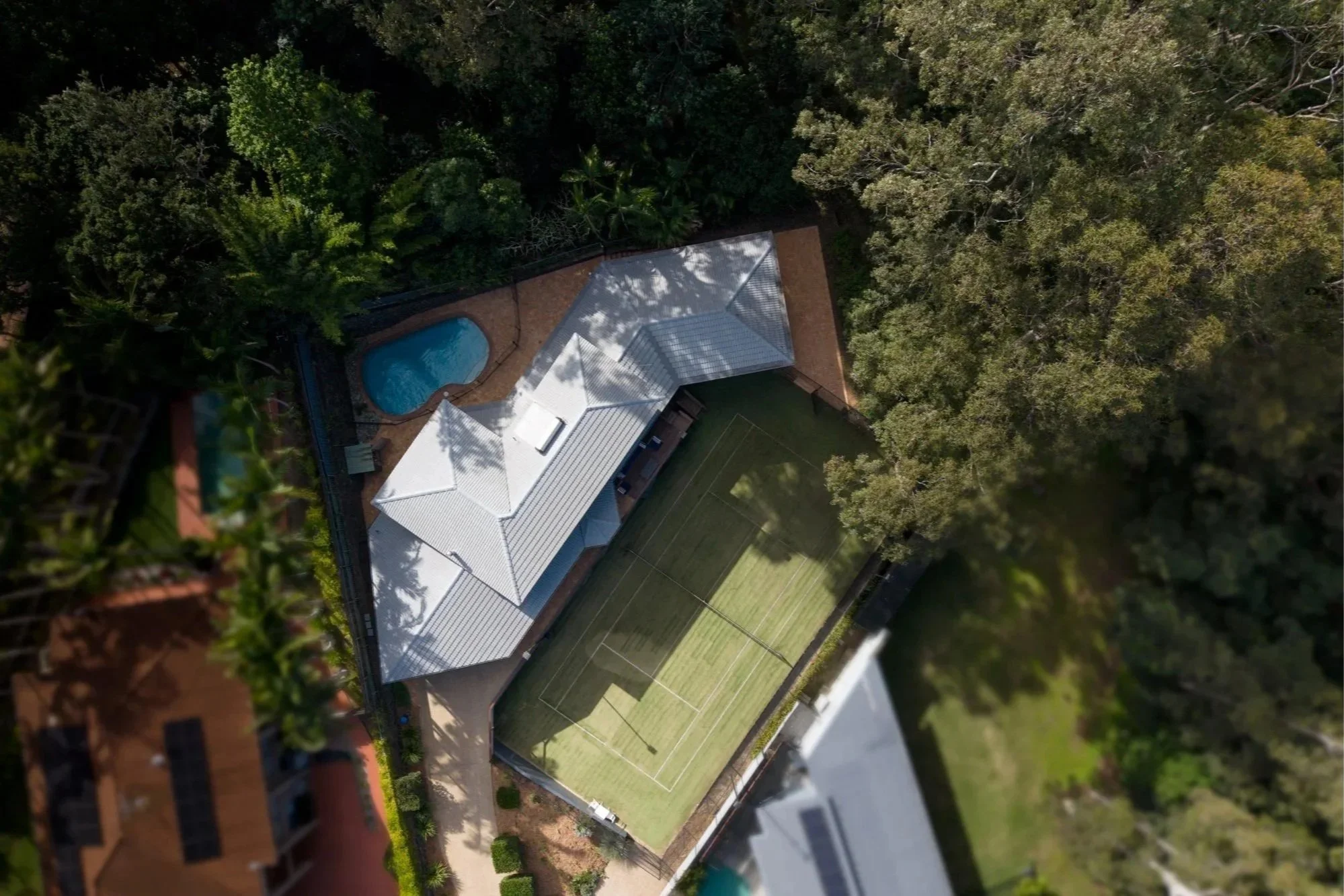 An aerial view of a backyard with a swimming pool, a tennis court, and a house surrounded by tall trees.