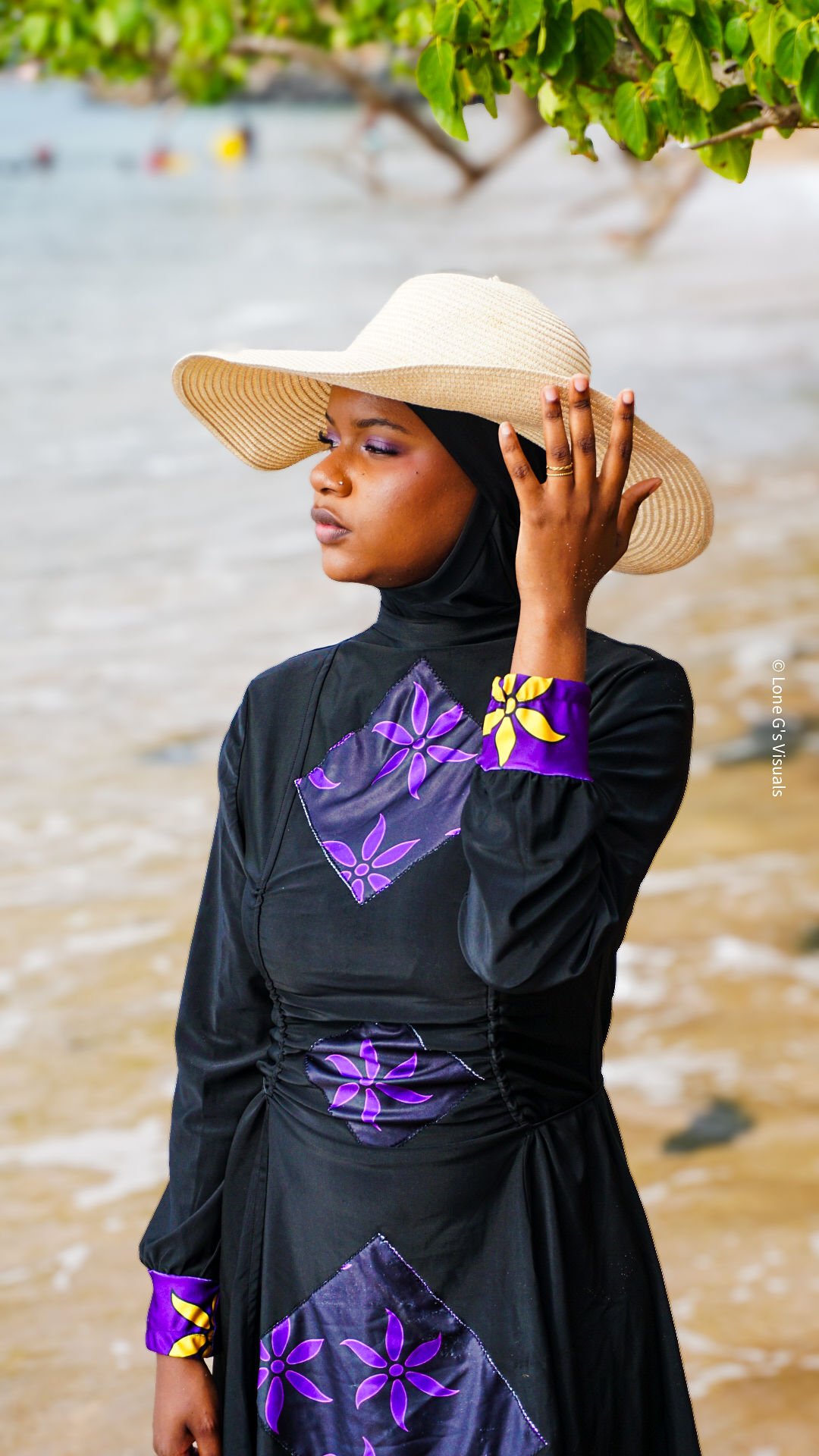 Une femme portant un chapeau de soleil et un burkini noir avec des motifs ylang violets et jaunes, se tenant près de la plage avec des eaux et des arbres en arrière-plan.