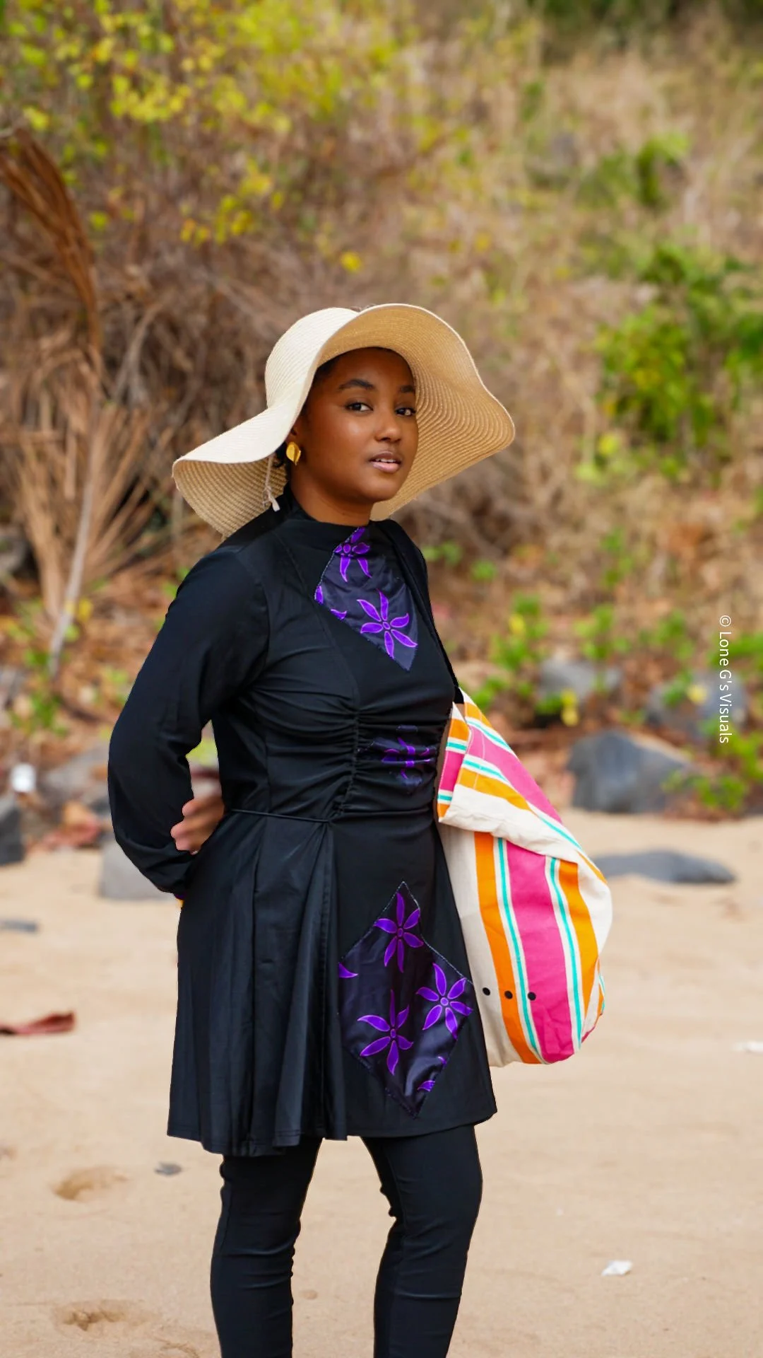Une femme portant un chapeau large, un burkini avec des motifs ylang violets, tenant un sac à main coloré, en bord de plage avec des rochers et des feuillages en arrière-plan.