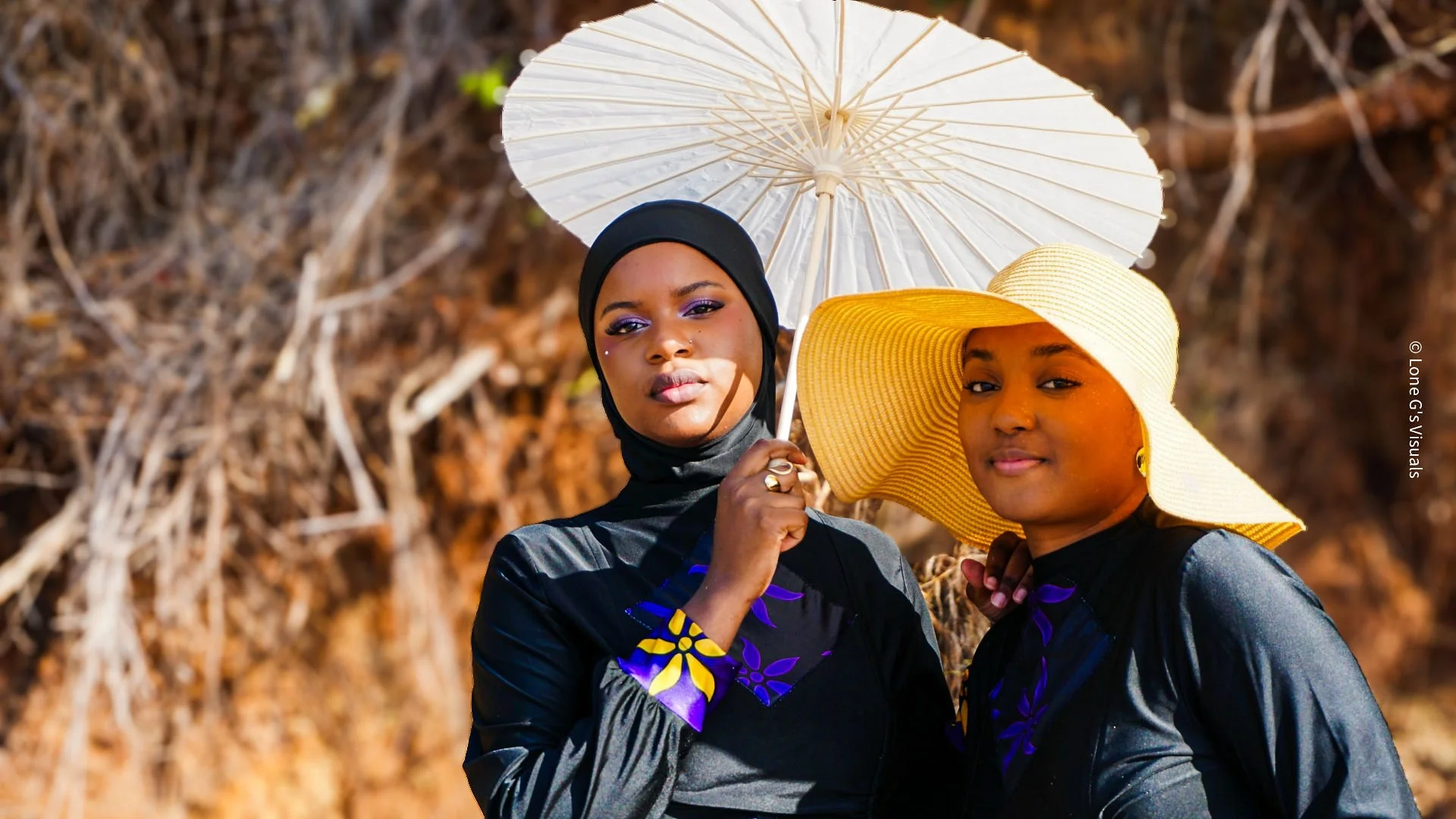 Deux femmes debout dans un environnement extérieur, tenant un parasol blanc. L'une porte un chapeau large jaune, l'autre un hijab noir. Elles portent toutes deux des burkinis noirs avec des motifs ylang violets et jaunes.