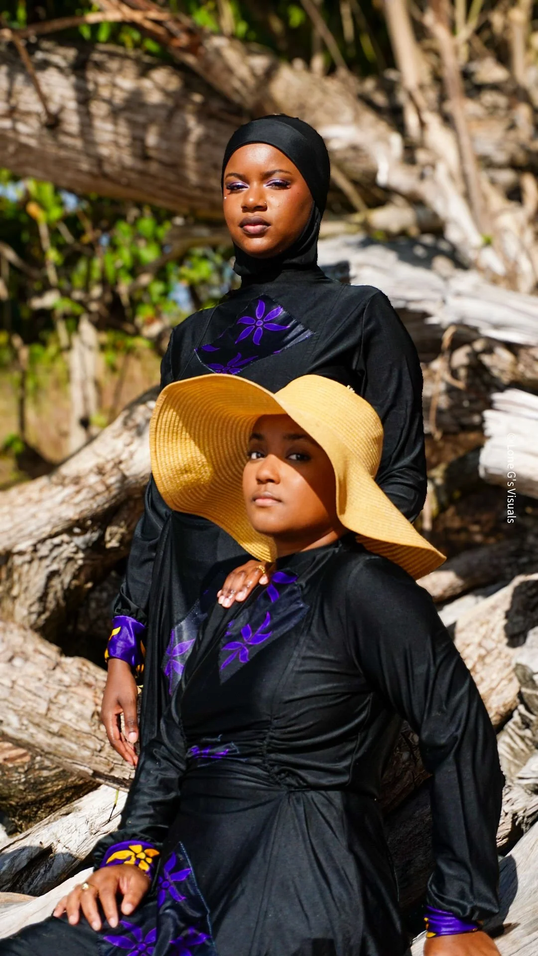 Deux femmes portent des burkinis noirs avec des motifs ylang violets, l'une porte un chapeau de paille et l'autre un bandeau noir, debout devant un fond de bois.