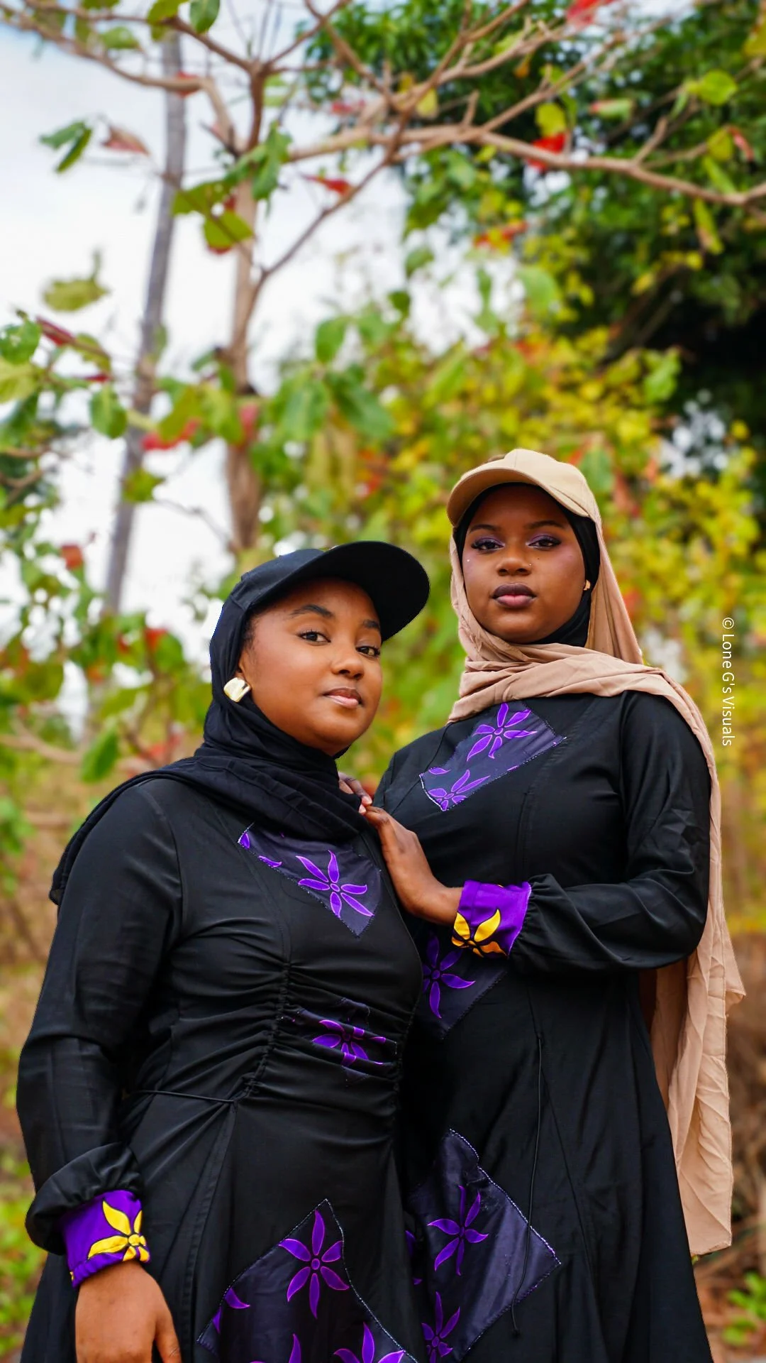 Deux jeunes femmes portant un burkini noir avec des motifs ylang violets, posant ensemble à l'extérieur dans un environnement naturel avec des arbustes en arrière-plan.