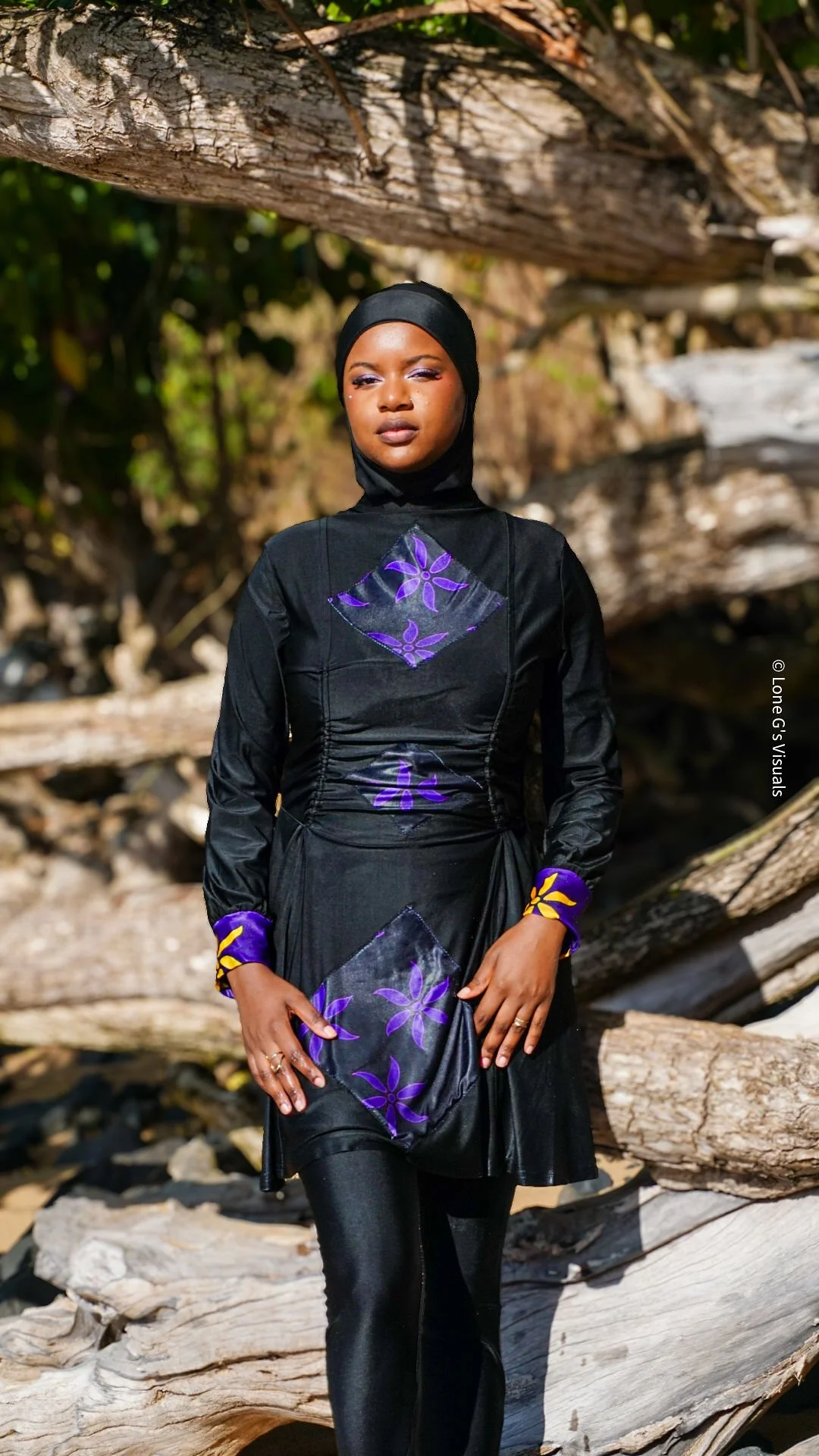Une femme portant un burkini noir avec des motifs ylang violets et jaunes, un foulard noir, se tient devant un bois flotté sur la plage.