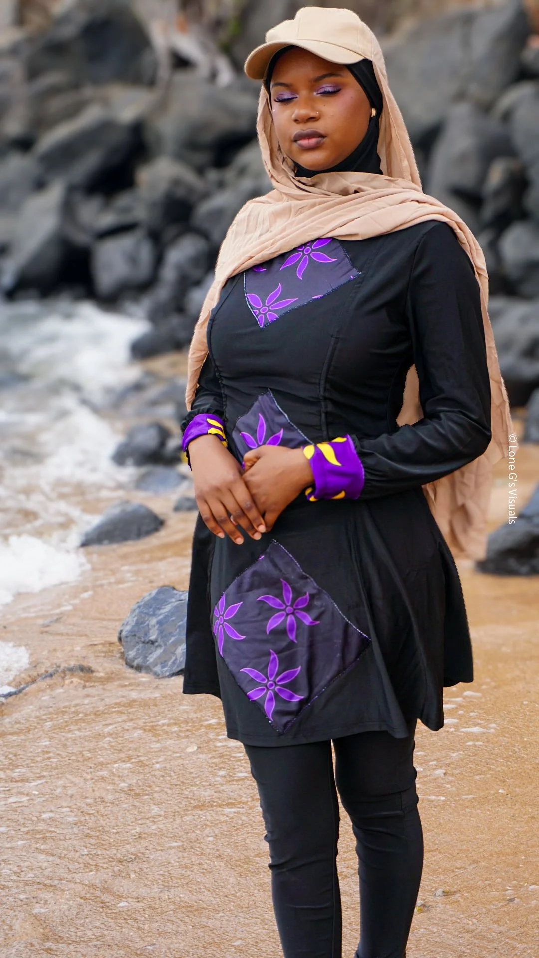 Une femme vêtue d'un burkini noir avec des motifs ylang violets et jaunes, portant une casquette avec voile intégré beige, se tient sur la plage avec des rochers autour.