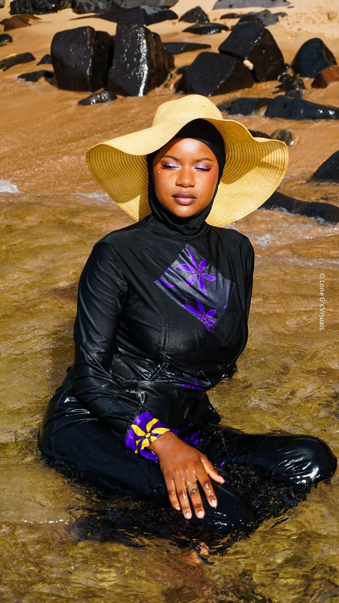Femme portant un chapeau en paille et un burkini noir avec des motifs ylang violets, assise dans l'eau avec des rochers noirs en arrière-plan.
