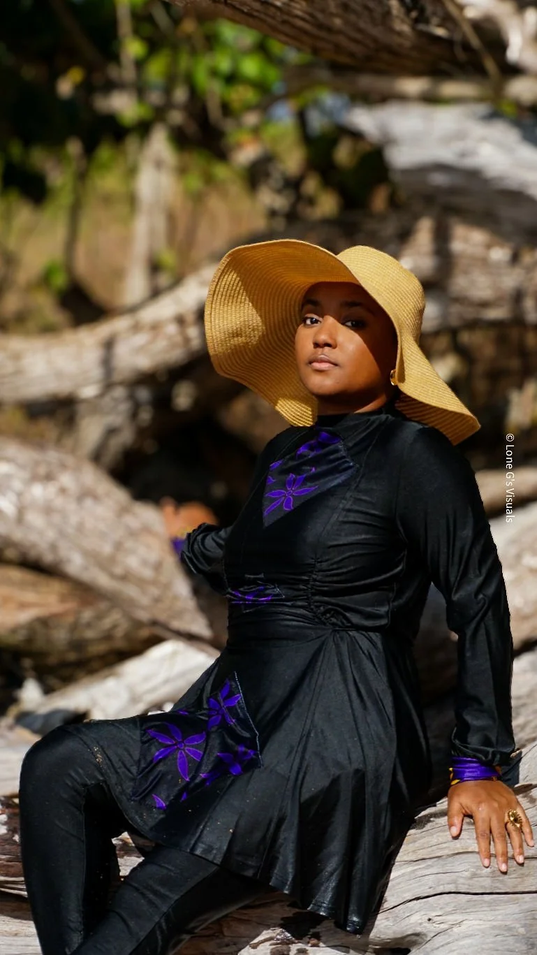 Femme portant un chapeau élégant assise sur un bois flotté, en arrière-plan des bois et des rochers, en plein air. La femme porte un burkini noir avec des motifs ylang violets.