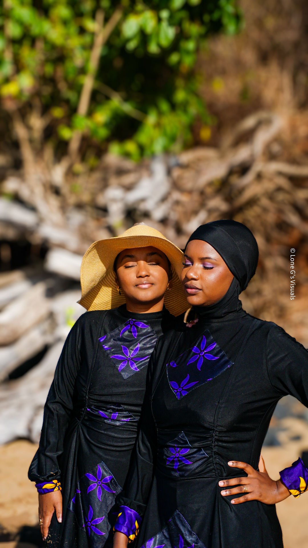 Deux femmes debout près d'une plage, portant des burkinis noirs avec des motifs violets, l'une avec un chapeau en paille et l'autre avec un foulard noir.