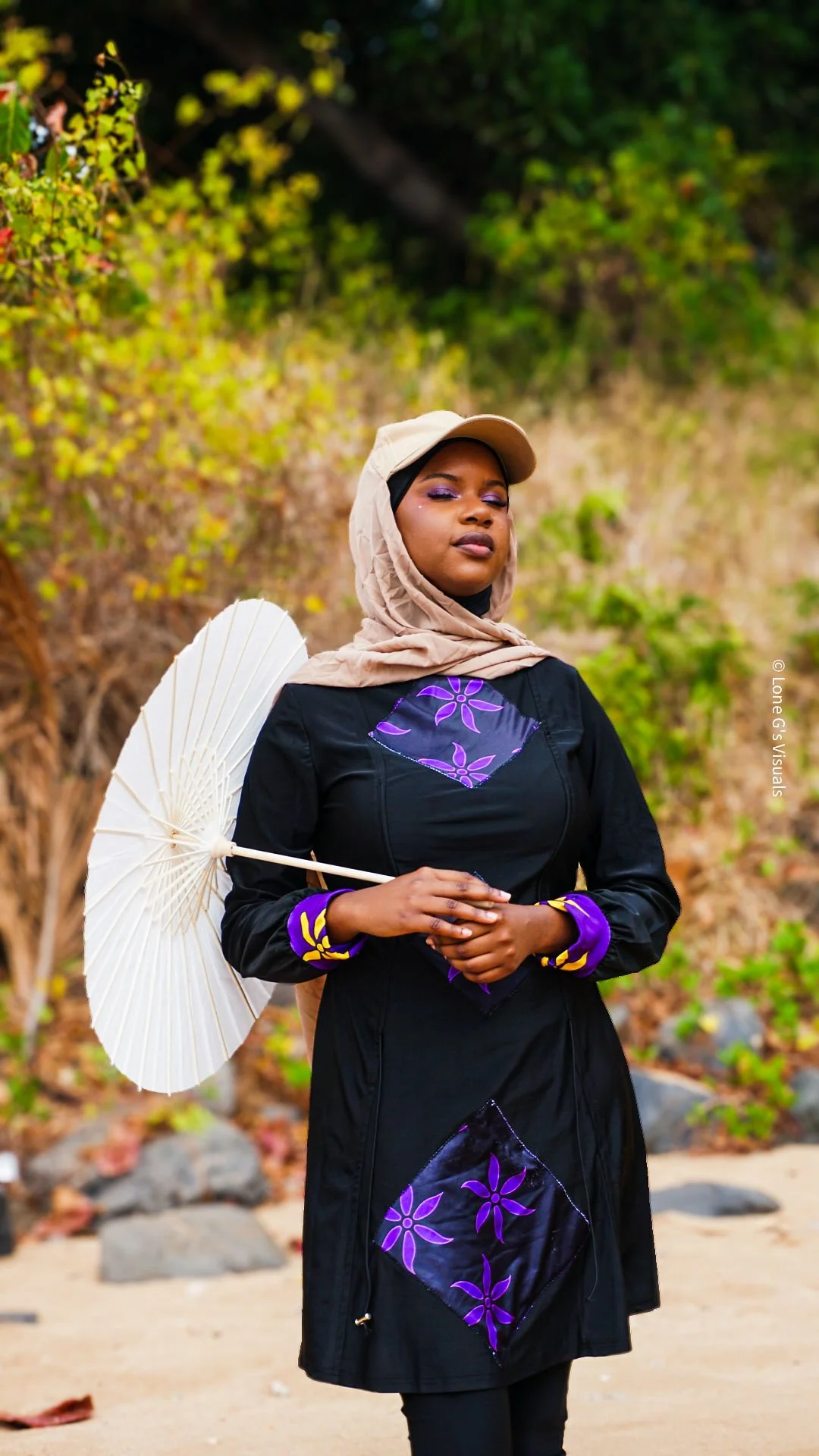 Jeune femme portant un burkini avec des motifs ylang violets, un foulard beige, une casquette avec voile intégré beige et tenant un parapluie blanc, en extérieur avec des arbres verts en arrière-plan.