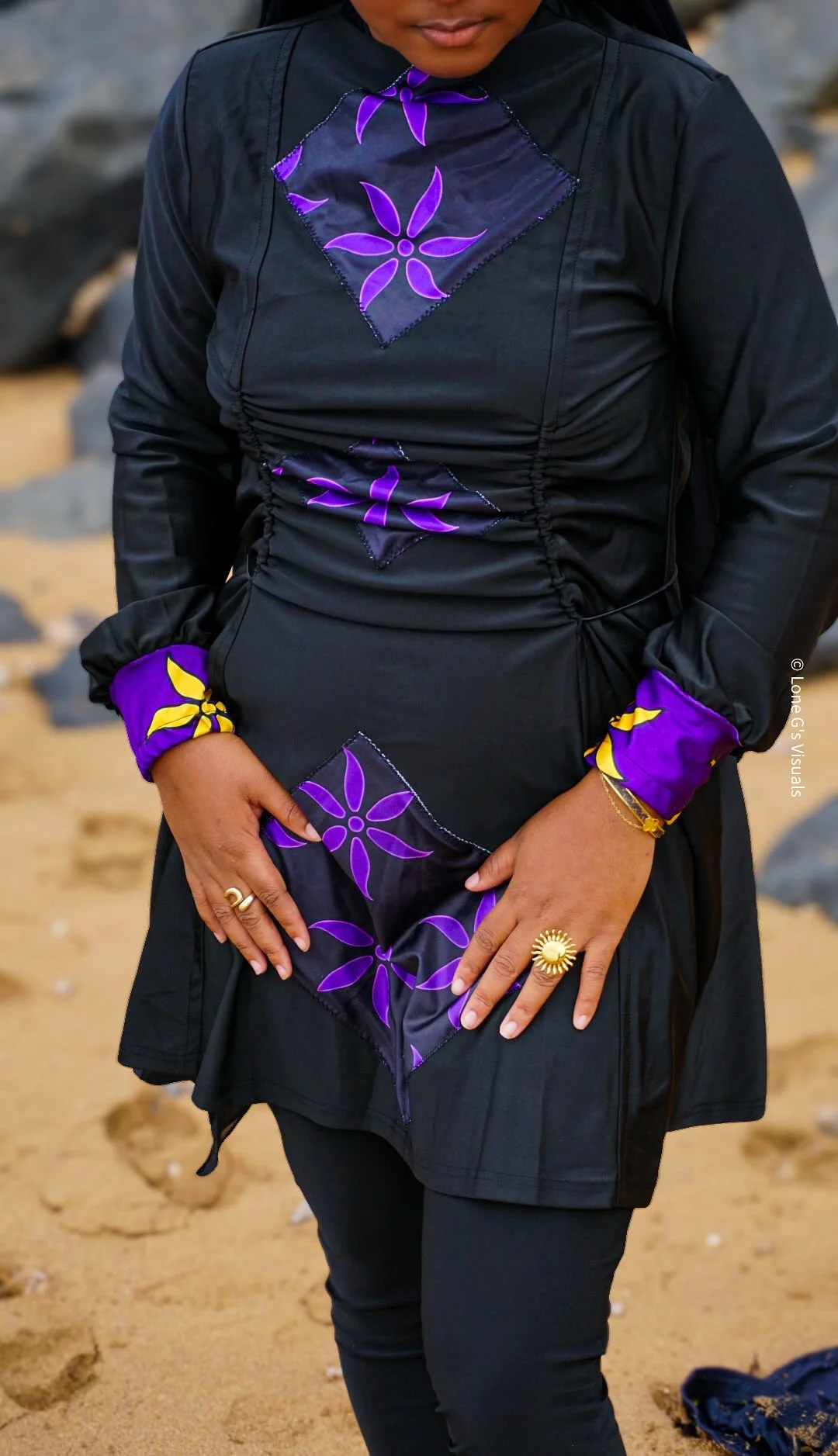 Une personne portant un burkini noir avec des motifs ylang violets et jaunes, avec des accessoires dorés, sur une plage.