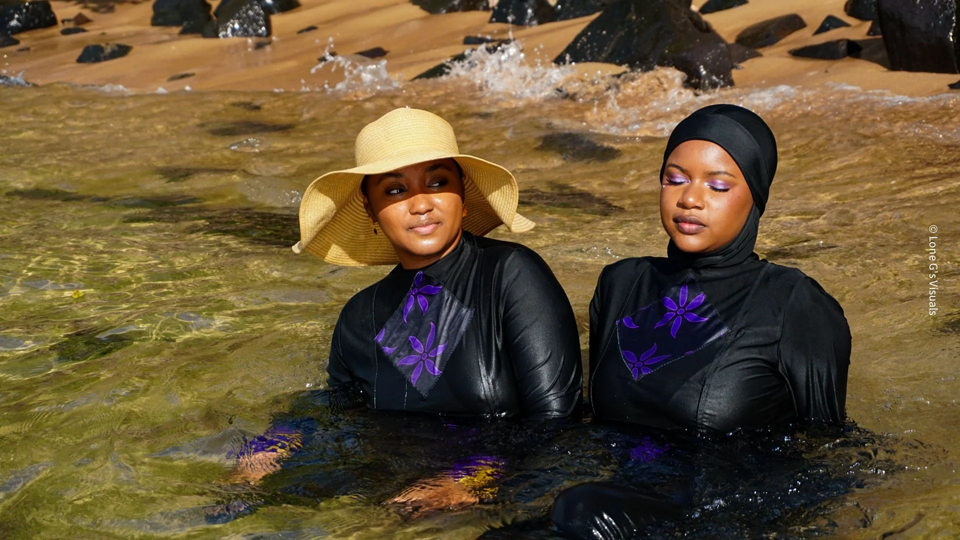 Deux femmes assises en bord de mer, portant des burkinis noirs avec des motifs ylang violets. L'une porte un chapeau en paille, l'autre un foulard noir, toutes deux avec des maquillages aux paupières violettes pâles.