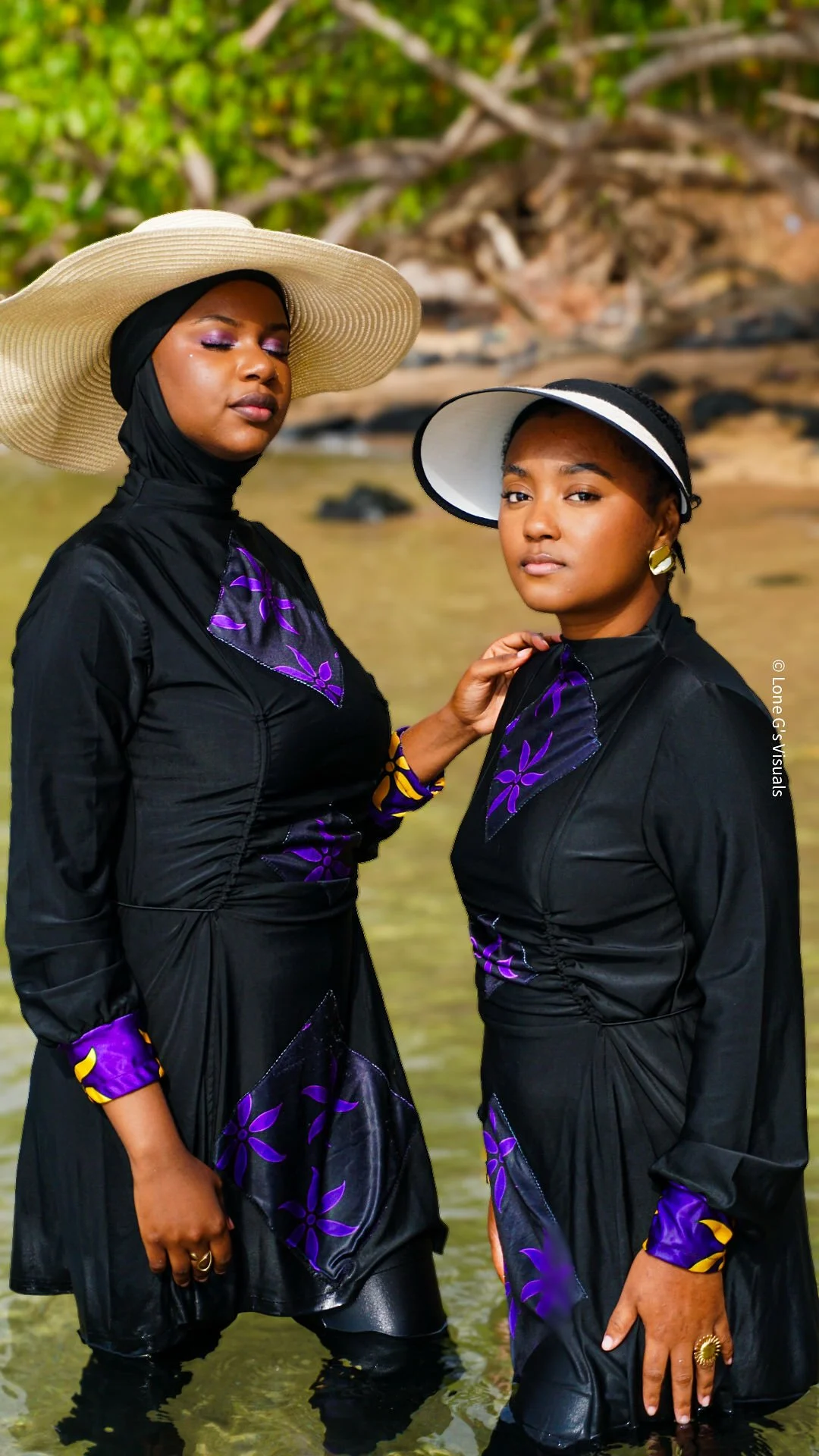 Deux femmes debout en bord de plage, portant des burkinis noirs avec des motifs ylang violets, larges chapeaux et des accessoires dorés, avec des arbres en arrière-plan.