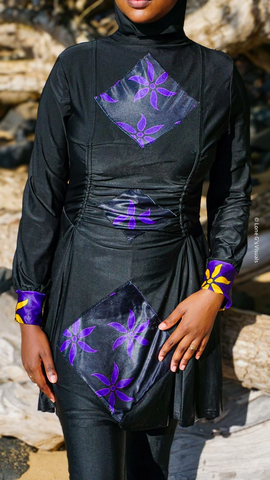 Une femme portant un burkini noir avec des motifs ylang violets et jaunes, près d'une plage rocheuse.