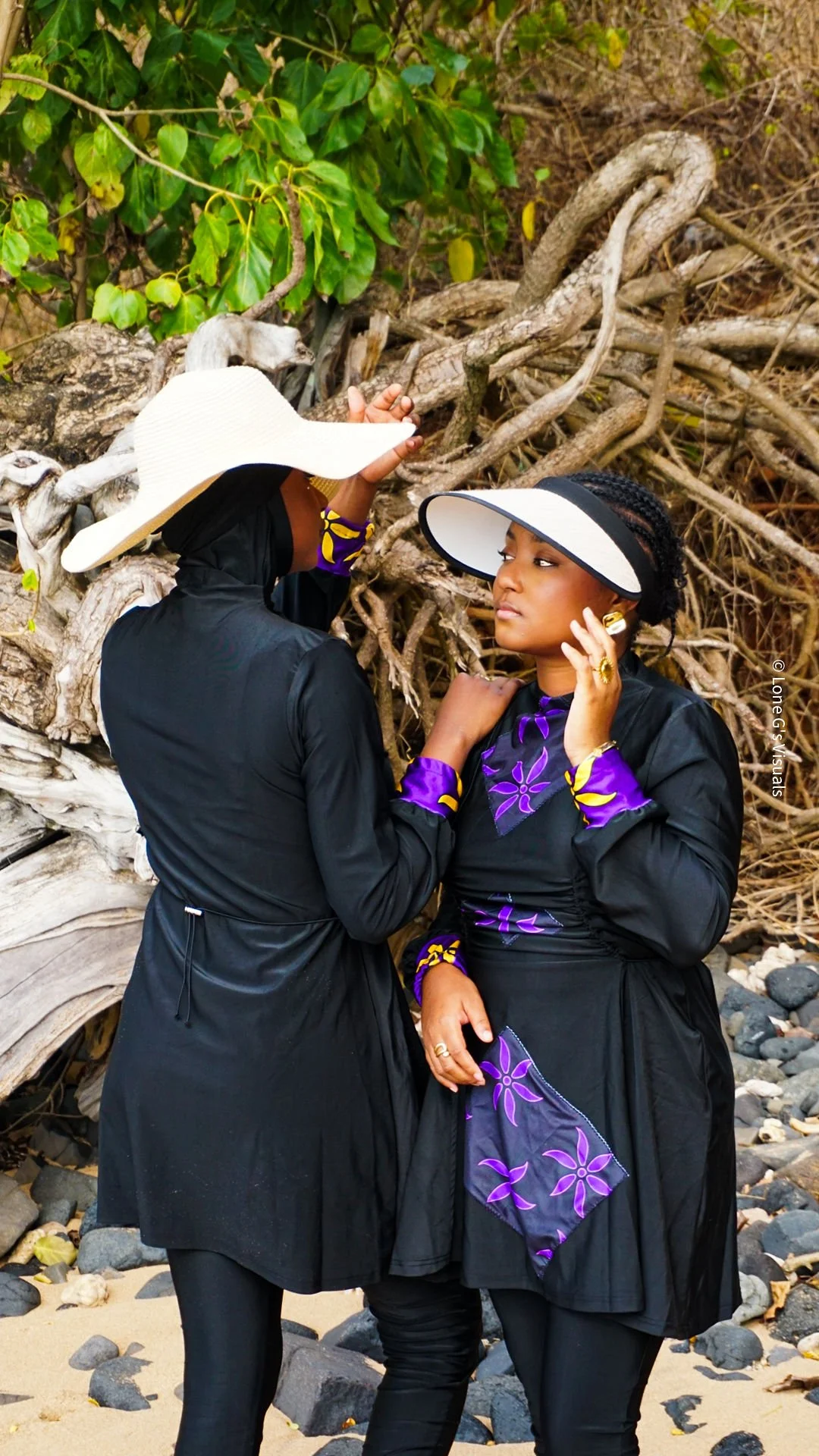 Deux femmes en tenue de plage et accessoires, se tiennent près d'une racine d'arbre sur une plage rocheuse.