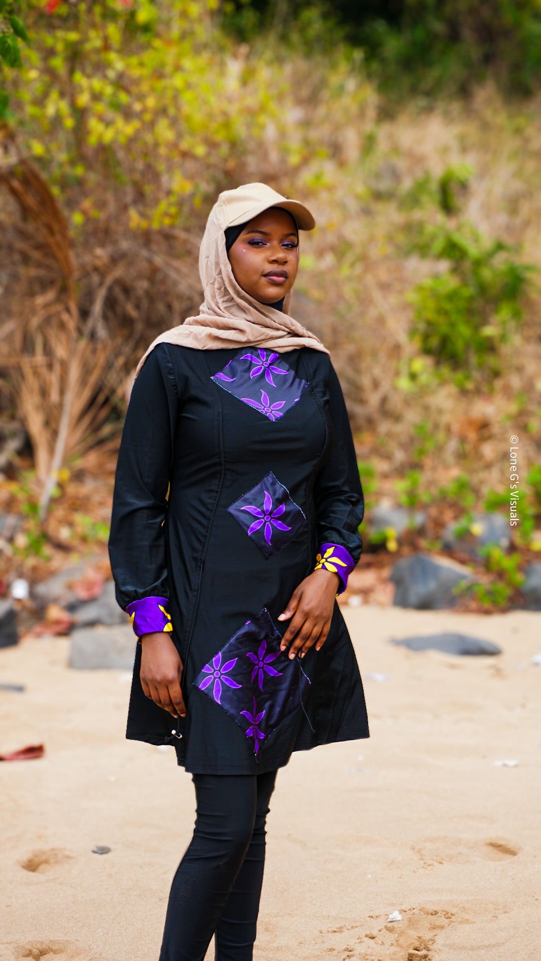 Jeune femme portant un burkini avec des motifs ylang violets, un foulard beige, une casquette avec voile intégré beige et se tient sur une plage avec des arbres en arrière-plan.