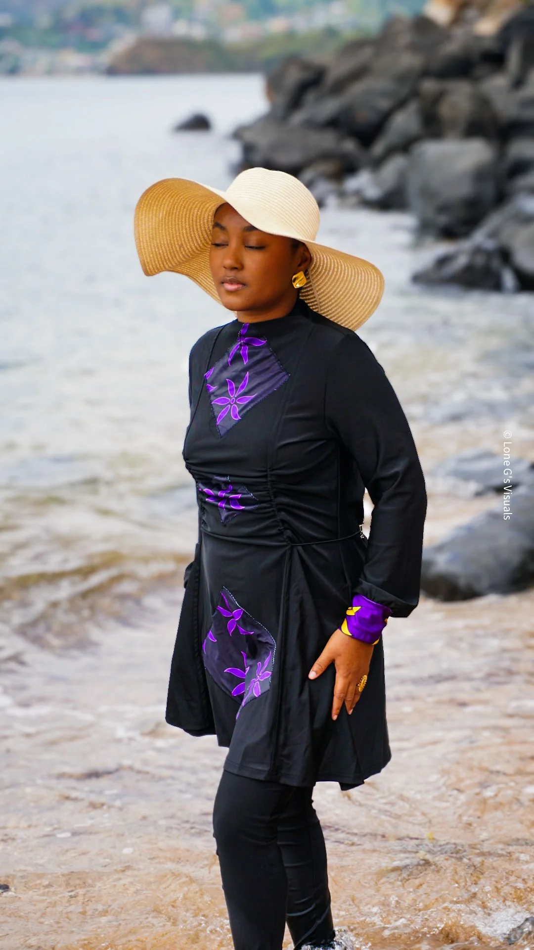 Jeune femme noire portant un chapeau de paille beige et un burkini avec des motifs ylang violets, se tenant sur la plage près de l'eau et des rochers, yeux fermés.