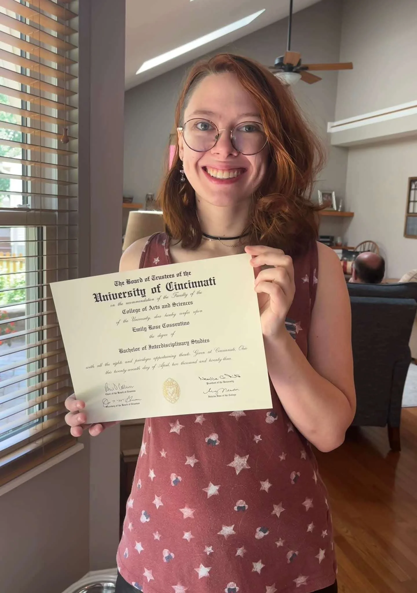 Emily is pictured with her red hair, glasses, and a big smile holding a graduation diploma, celebrating her graduation from the University of Cincinnati.
