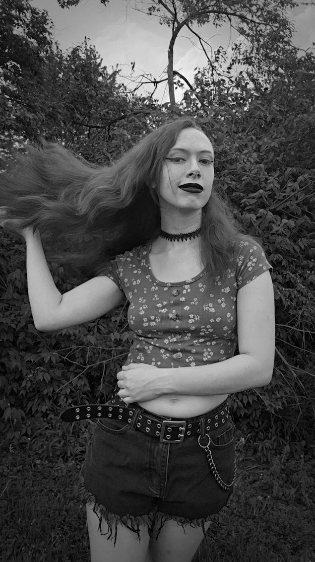 Emily poses confidently outdoors in front of dense bushes and trees, flipping her long hair to the side, wearing a floral crop top, a choker necklace, black shorts with a chain, and dark lipstick, captured in black and white.