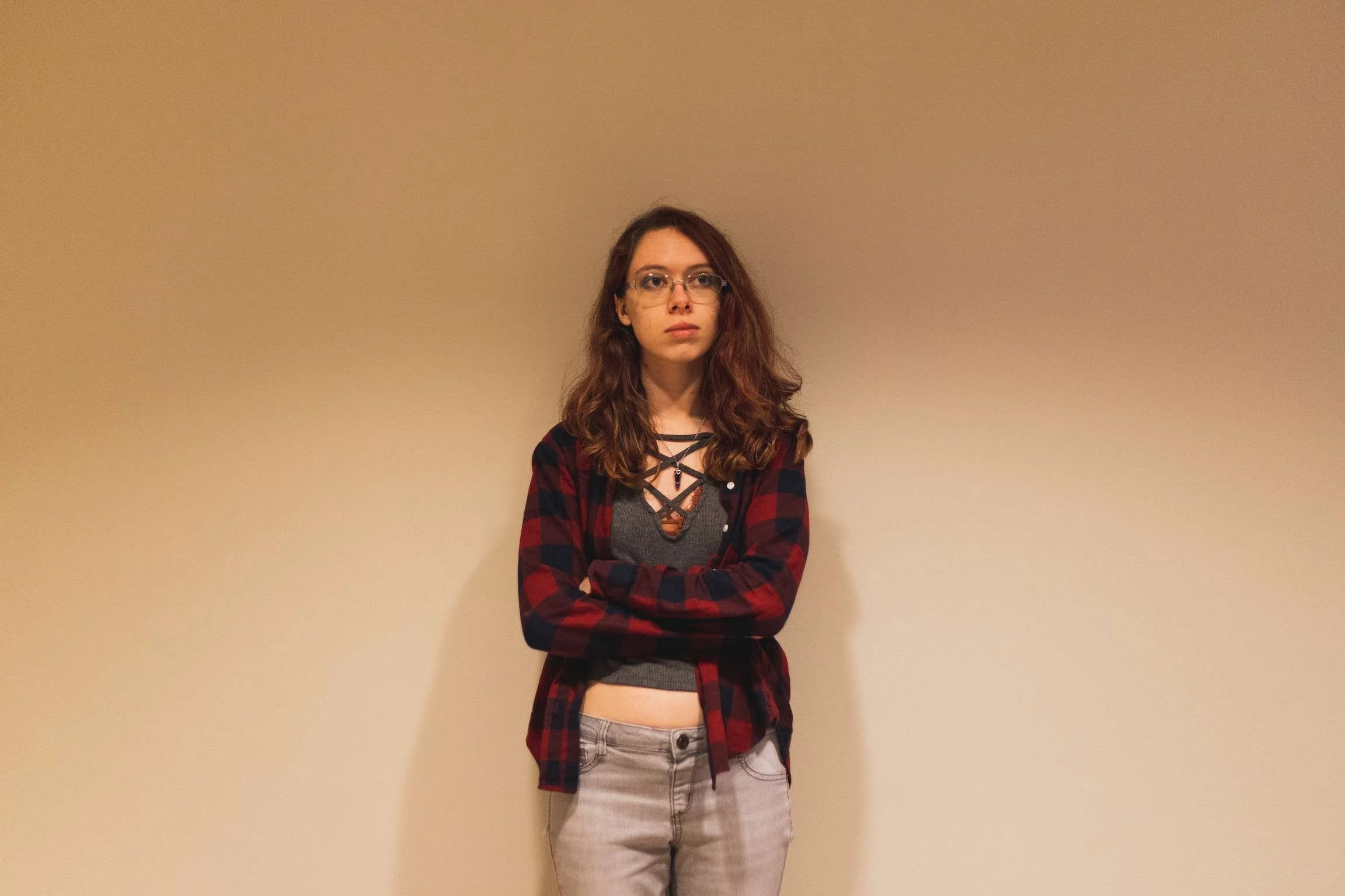 Emily poses, standing against a plain beige wall, wearing a red and black plaid shirt over a gray top, with her arms crossed.