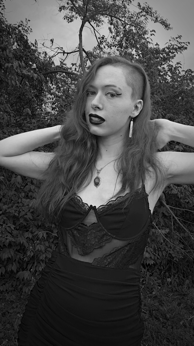Emily poses outdoors in front of trees and branches with her long wavy hair with one side shaved, wearing dark lipstick, earrings, and a pendant necklace.