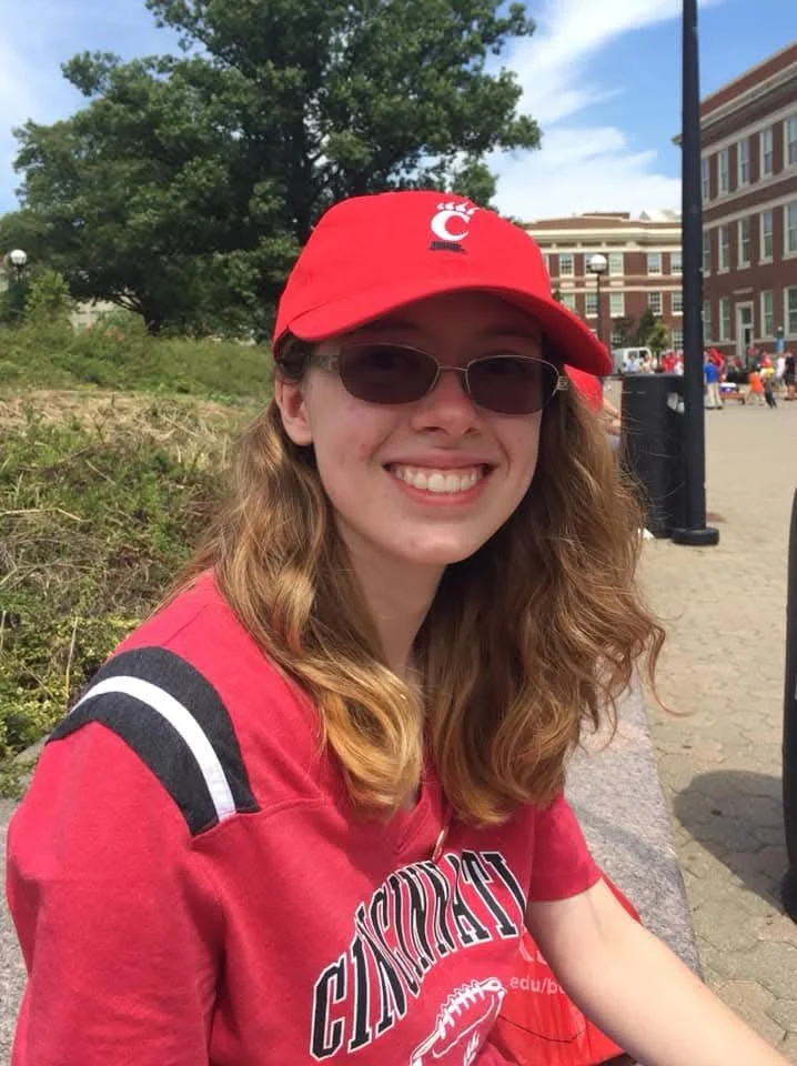 Emily sits on campus, wearing sunglasses, a red Cincinnati Reds baseball cap, and a red sports jersey, smiling on a sunny day.