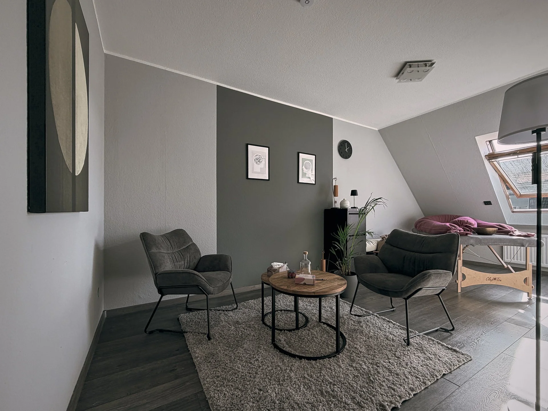 Moderner Wohnzimmerbereich mit zwei schwarzen Sesseln, einem runden Holztisch, Teppich, Wandkunst und Pflanzen, mit Bett an der Fensterseite.