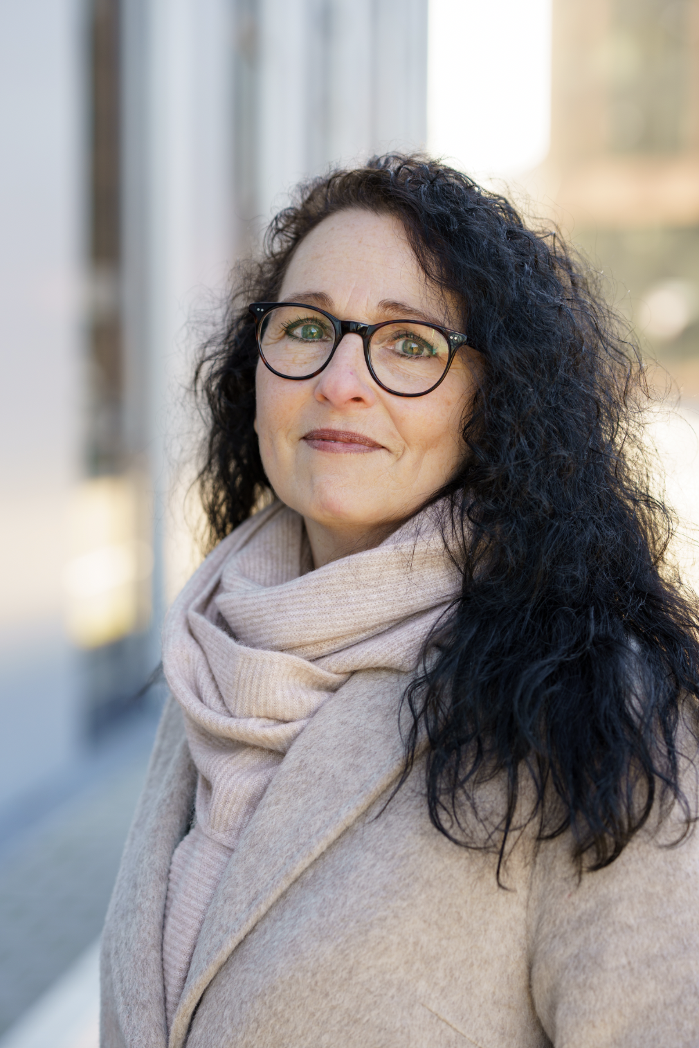 Eine Frau mit schwarzen, lockigen Haaren, Brille und einem beigen Mantel mit Schal steht draußen auf einer Straße, wahrscheinlich im Herbst oder Winter.