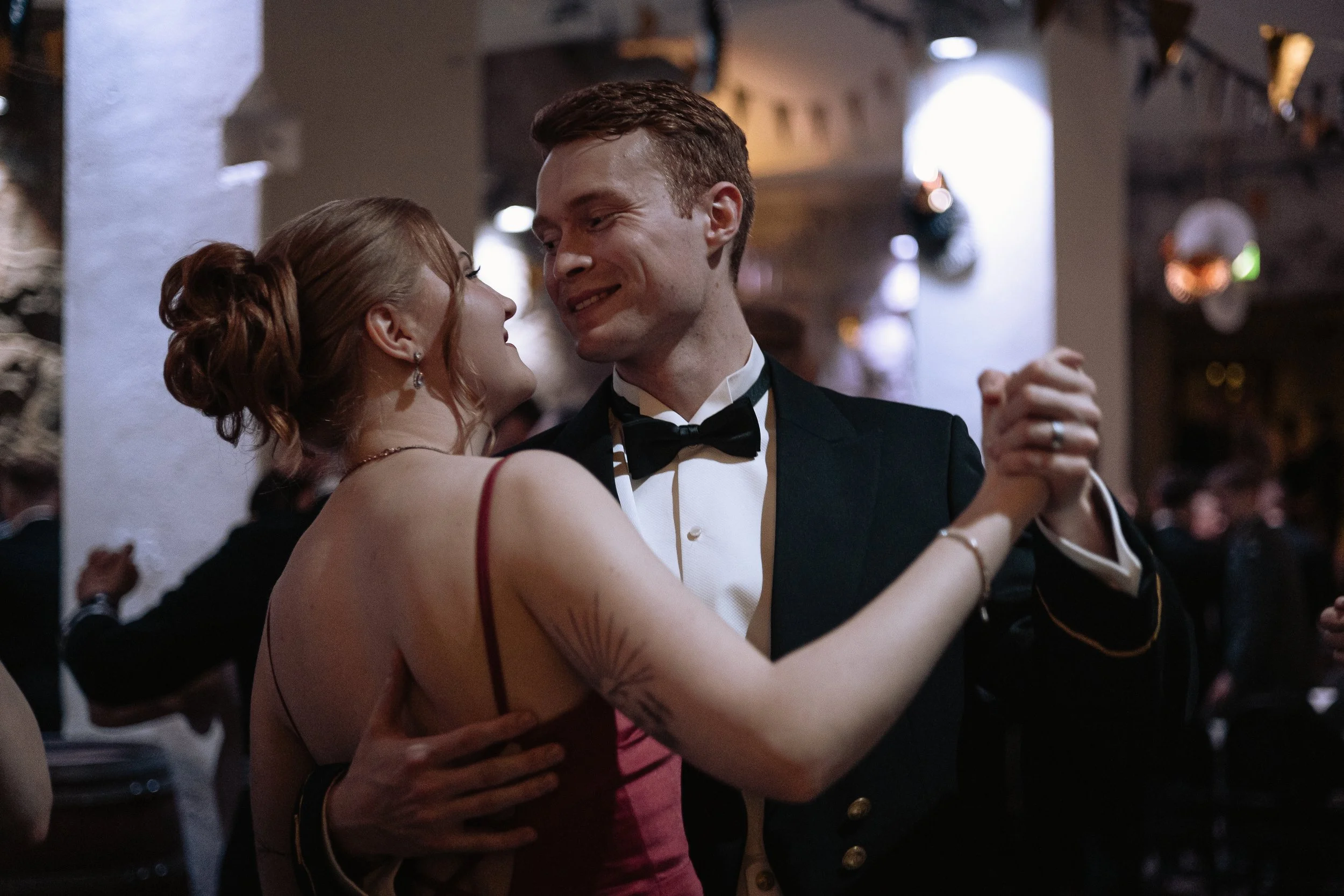 A man and woman dancing closely at a formal event, with the woman in a red dress and the man in a tuxedo, smiling and looking at each other.