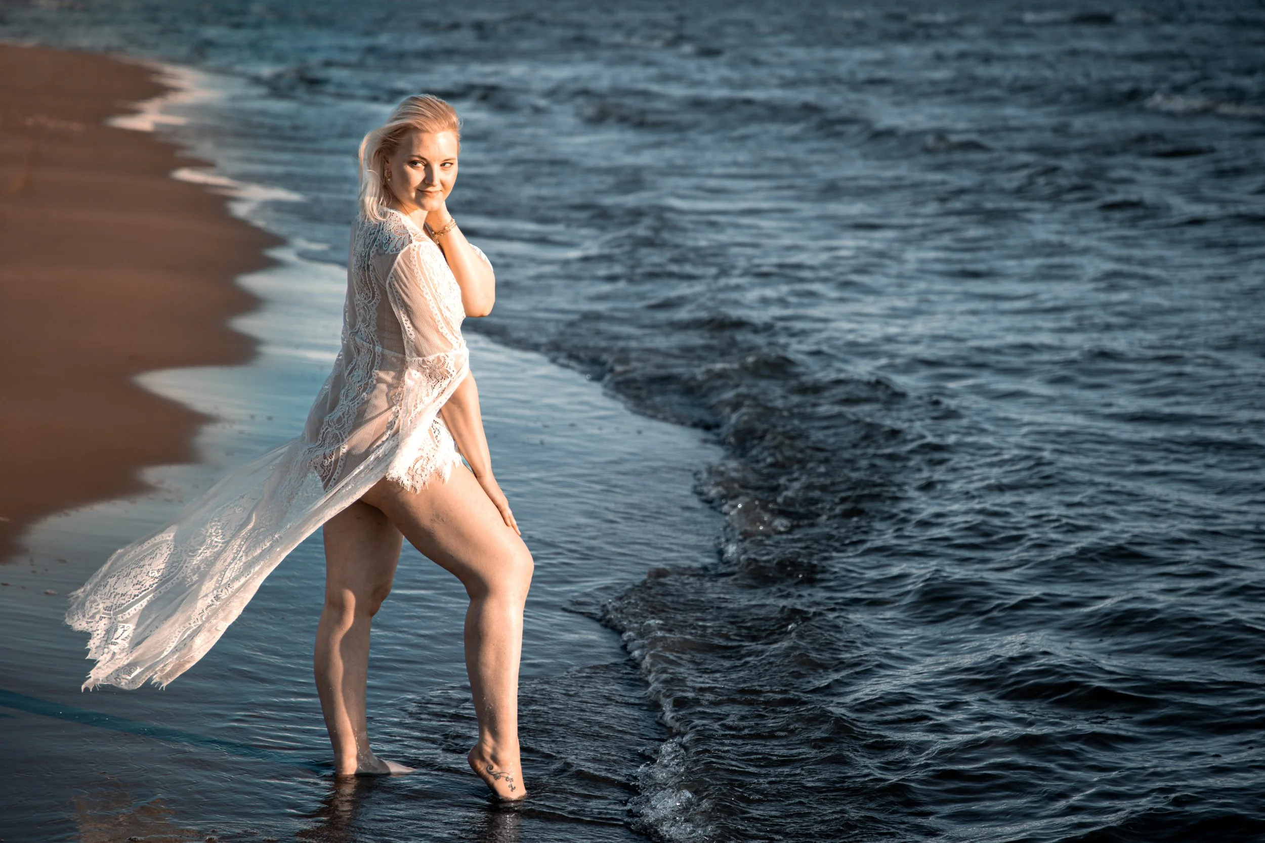 A woman with blonde hair standing on the beach at the edge of the water, wearing a see-through lace cover-up over her swimwear, with her hand on her chin, looking at the camera, during sunset or early evening.