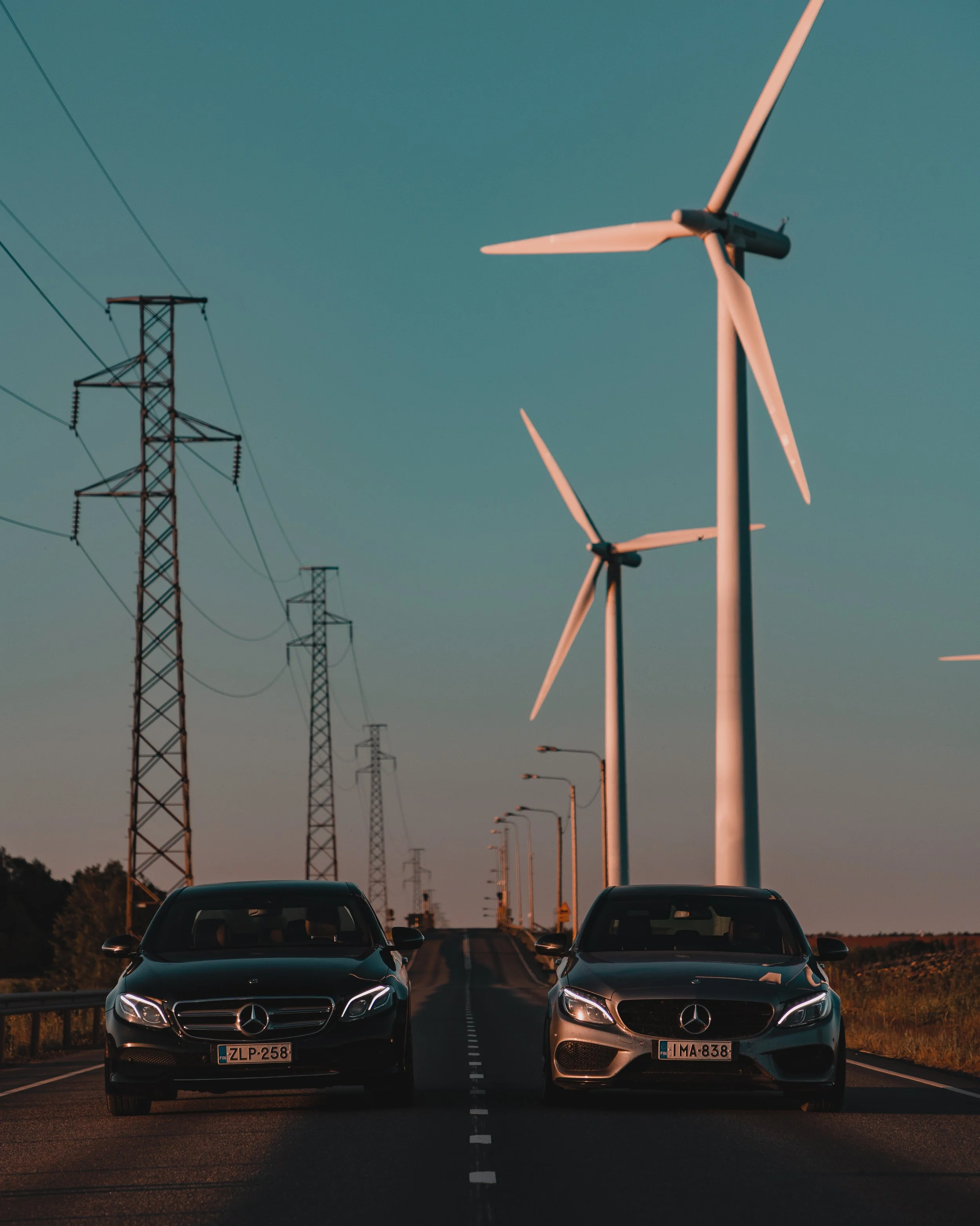 Two Mercedes-Benz cars parked side by side on a road with wind turbines and power lines in the background during sunset.