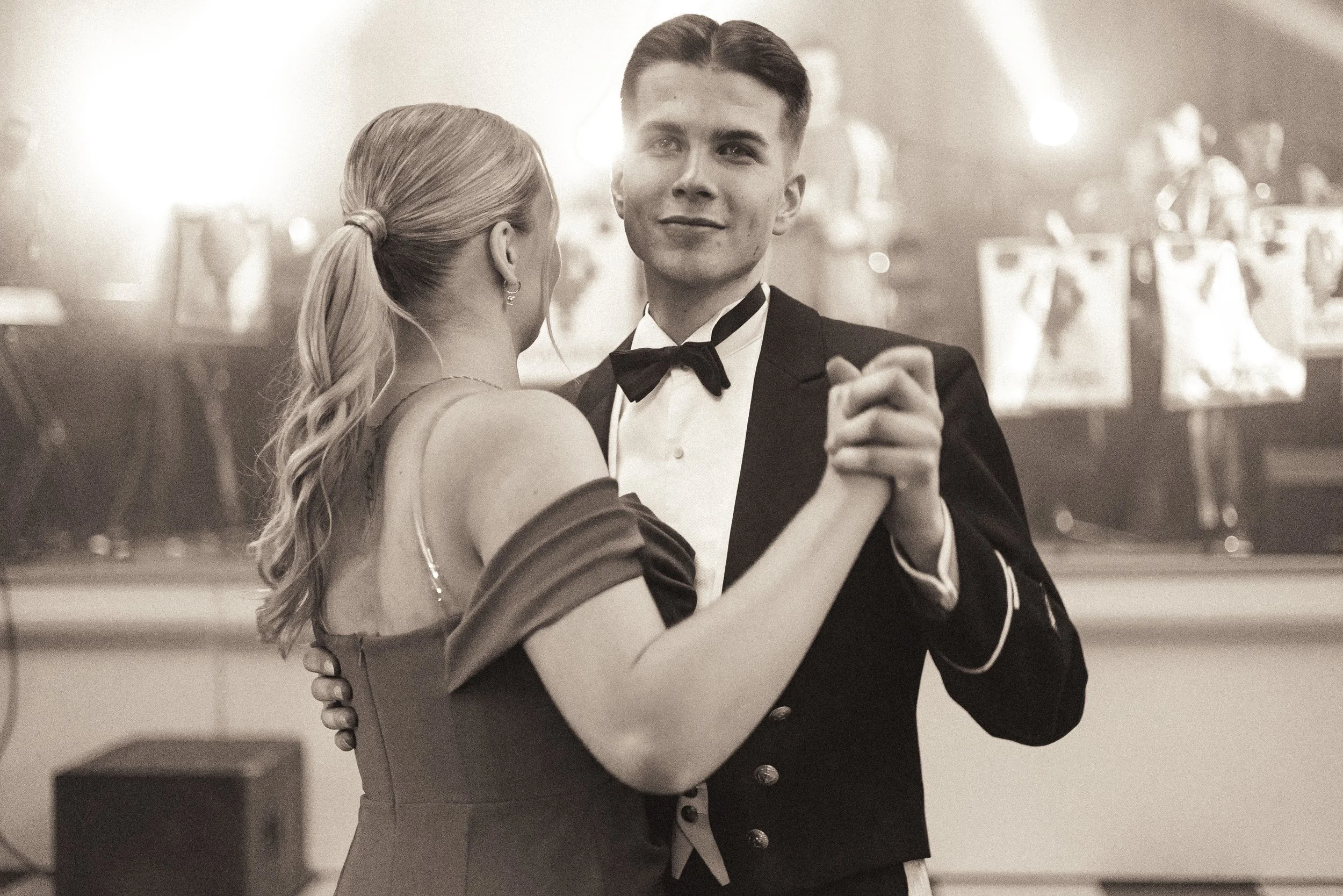 A young man and woman dancing at a formal event, dressed in evening attire, with the man wearing a tuxedo and the woman wearing an elegant dress, with a blurred background of the venue.