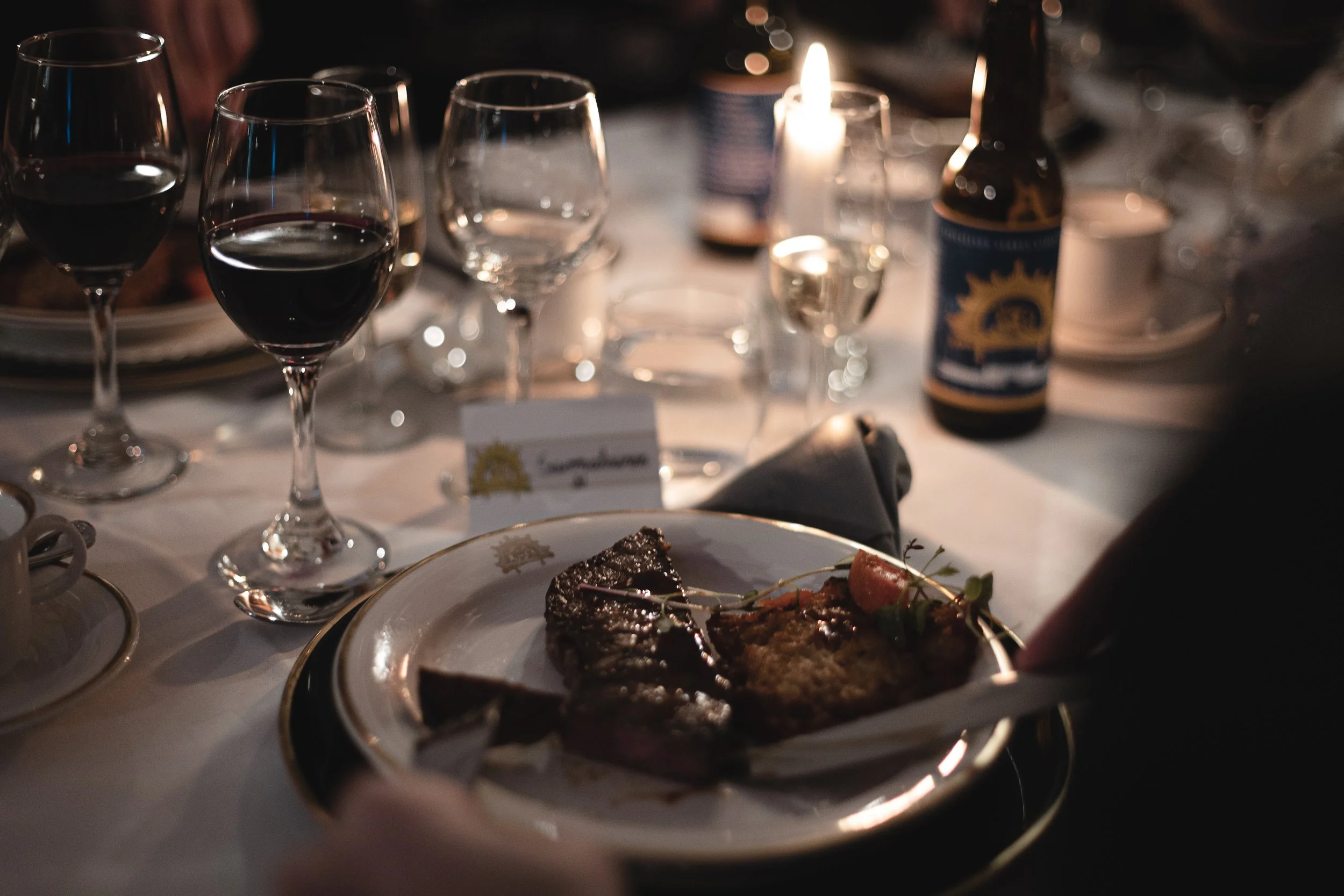 A dinner table set with multiple wine glasses, a bottle of beer, and a plate with steak and grilled vegetables, illuminated by candlelight.