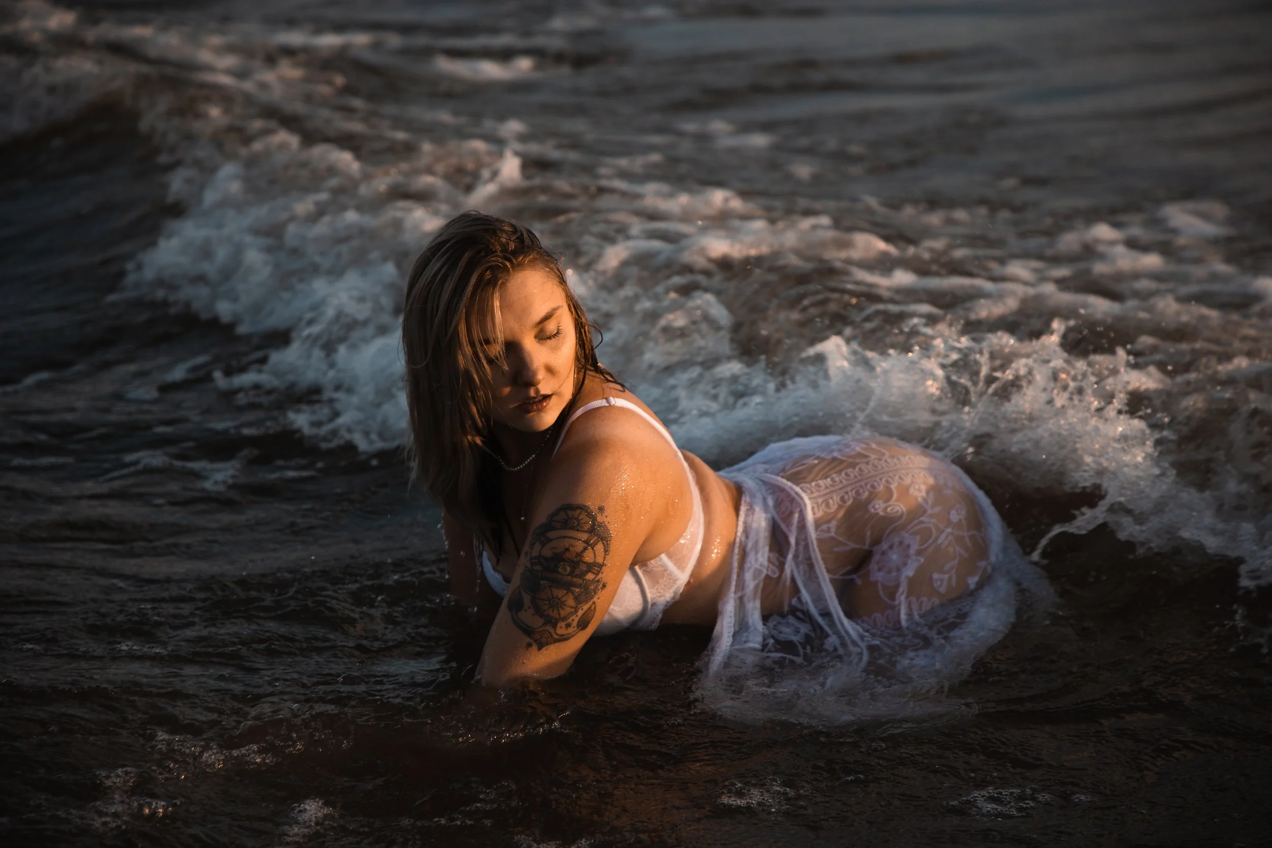 Woman with tattoo on her arm in white lace clothing, sitting in dark water on a beach at sunset.