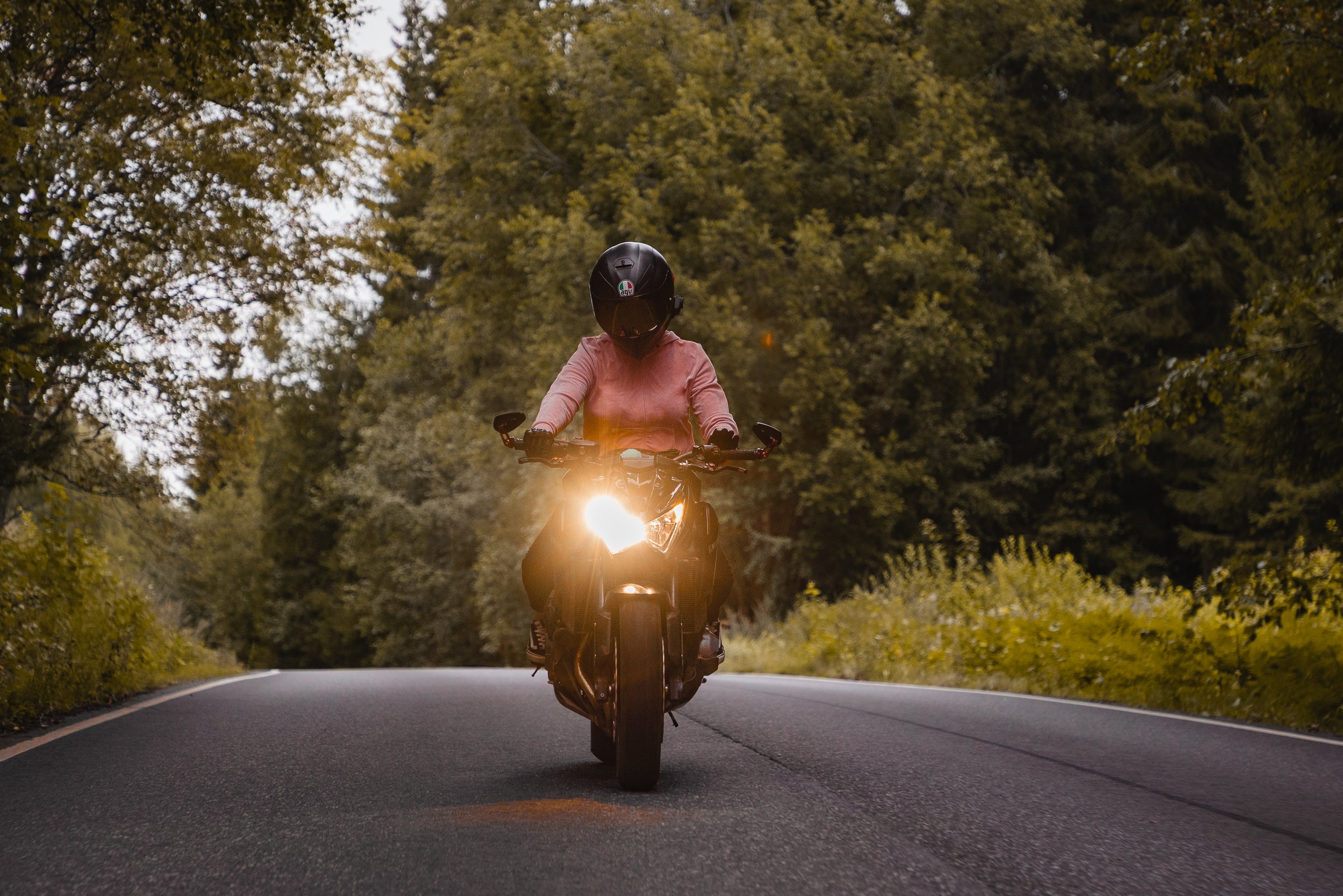 Person riding a motorcycle on a winding road surrounded by trees, with the motorcycle's headlight on, during sunset.