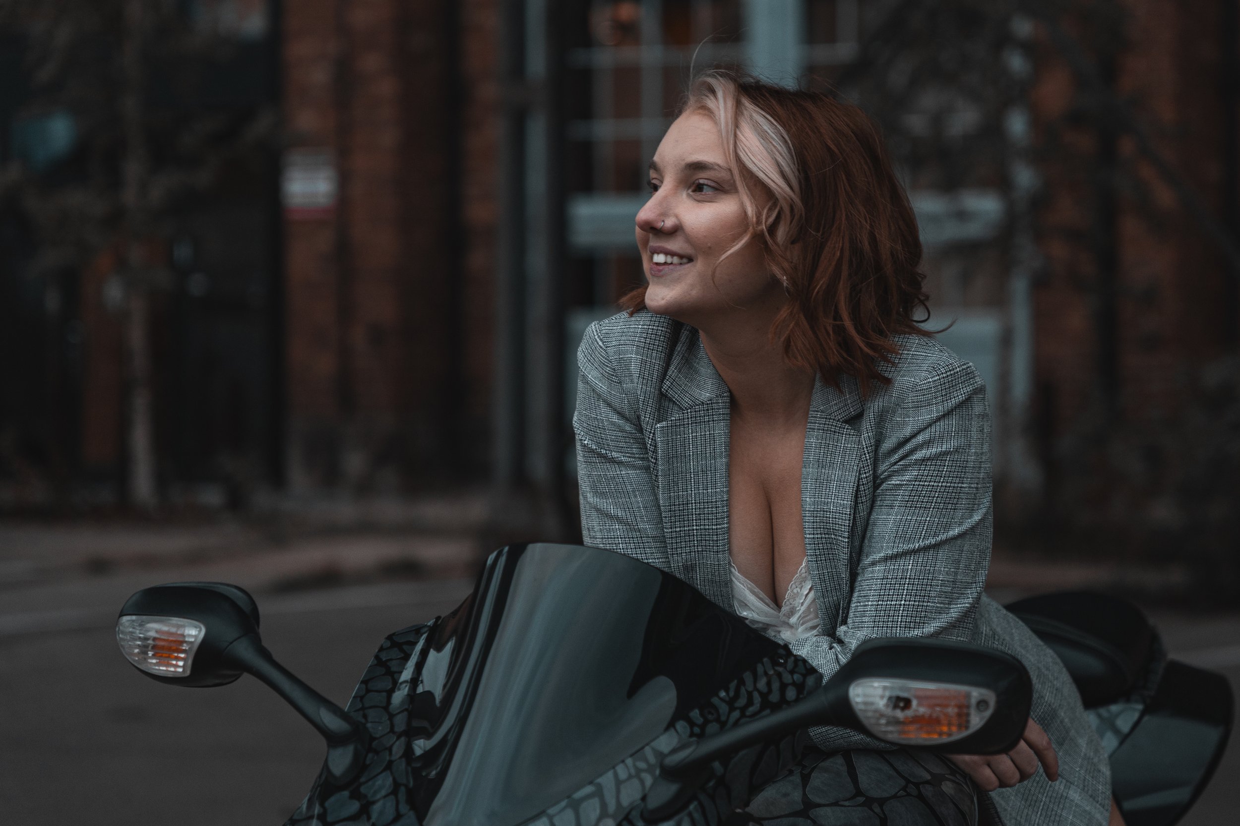 A woman with shoulder-length red hair and a nose piercing sitting on a motorcycle, smiling and looking to the side, in an urban outdoor setting during the evening or dusk.