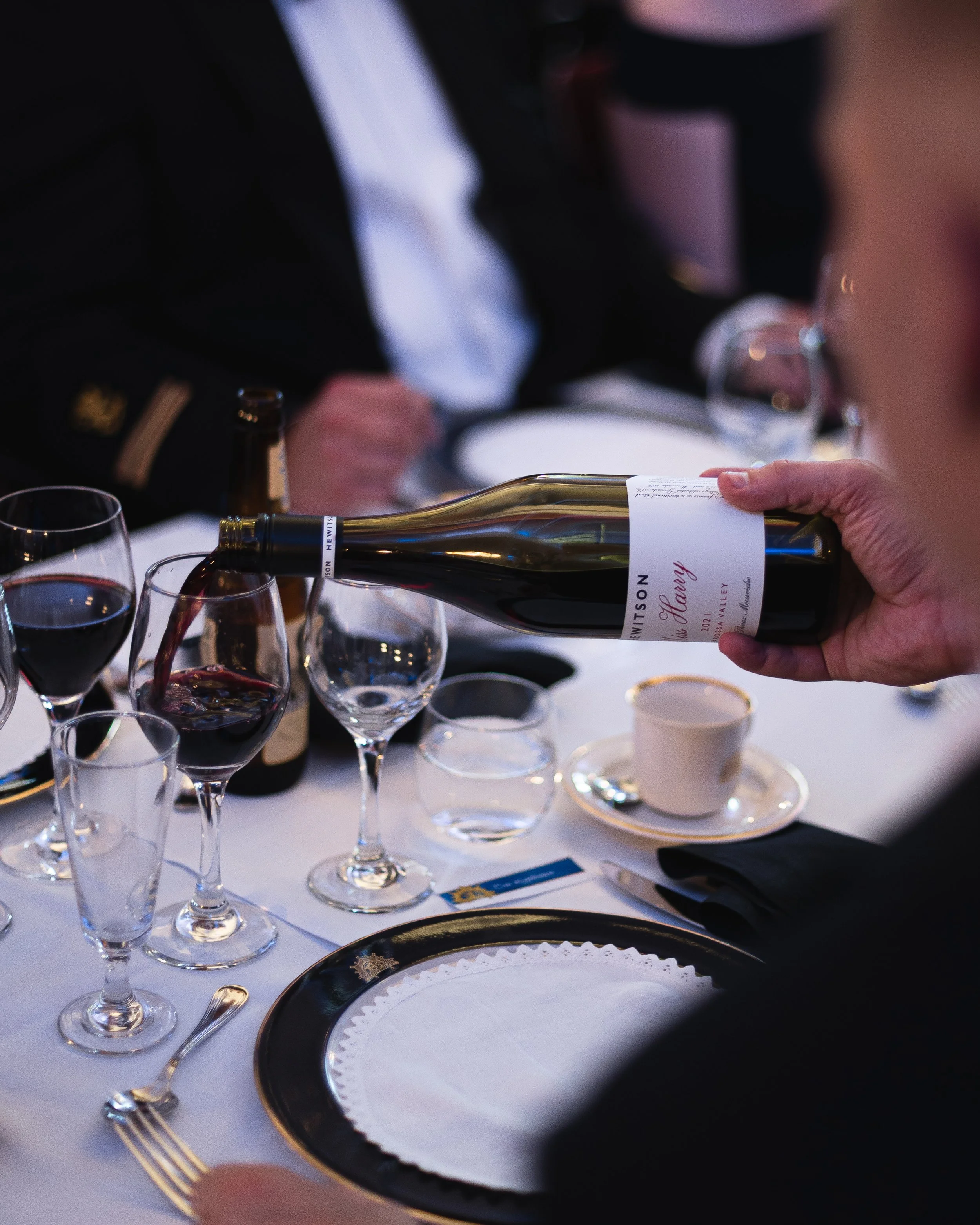 Person pouring red wine into a glass at a formal dinner table set with wine glasses, a cup and saucer, silverware, and a black and white plate.